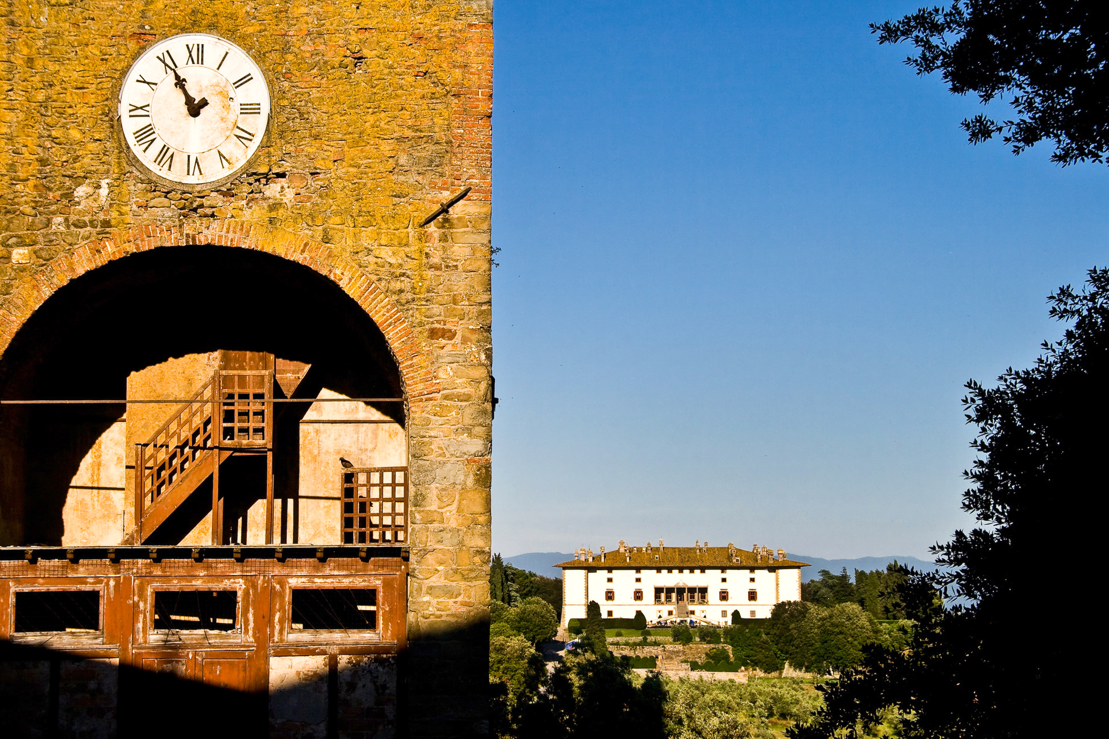 Clock tower on stone archway overlooking a grand Tuscan villa and landscape.