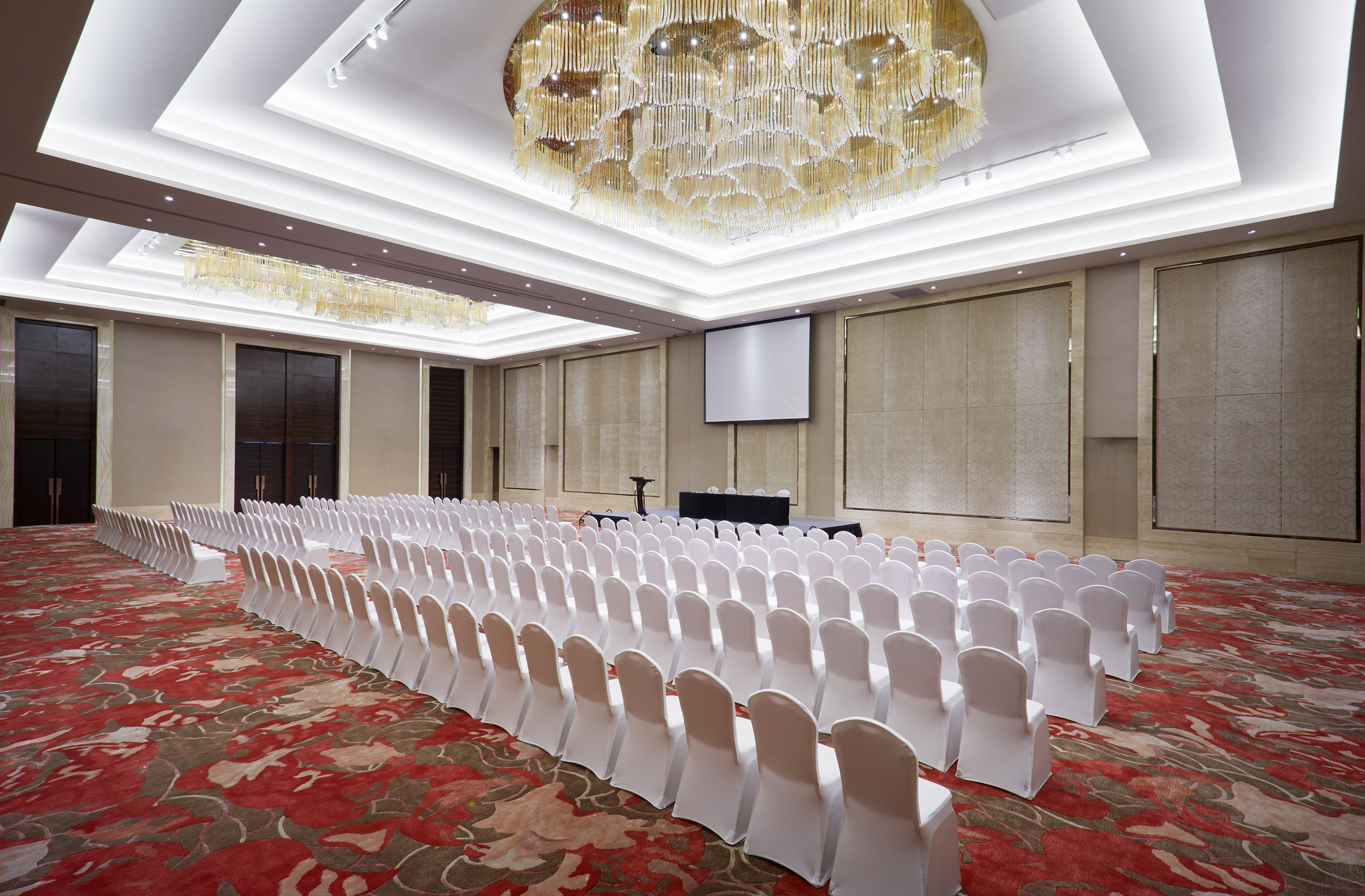 Empty conference ballroom with white chairs, red carpet, screen, and chandeliers.