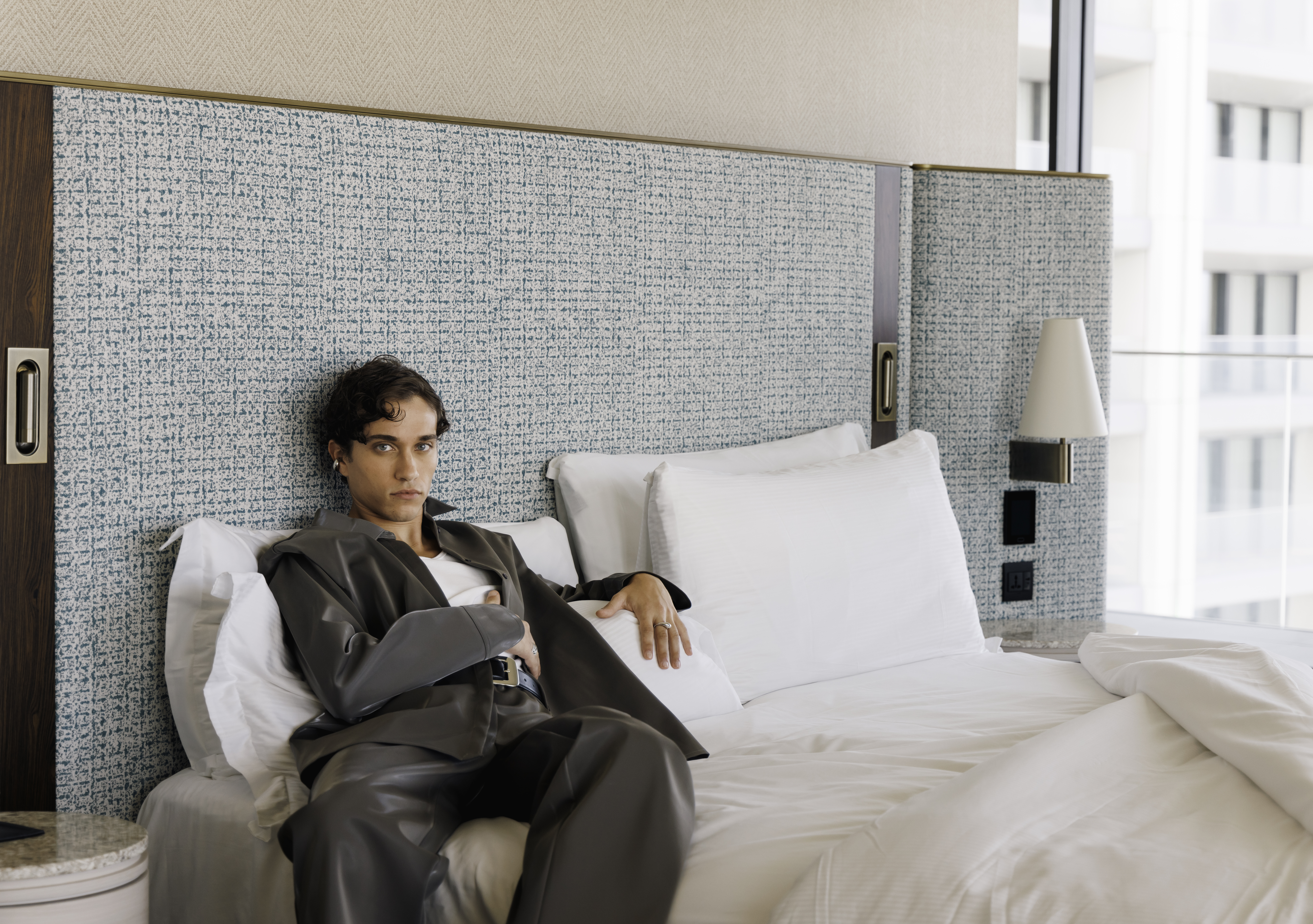 Stylish man with blue eyes in grey suit lounging on hotel bed.