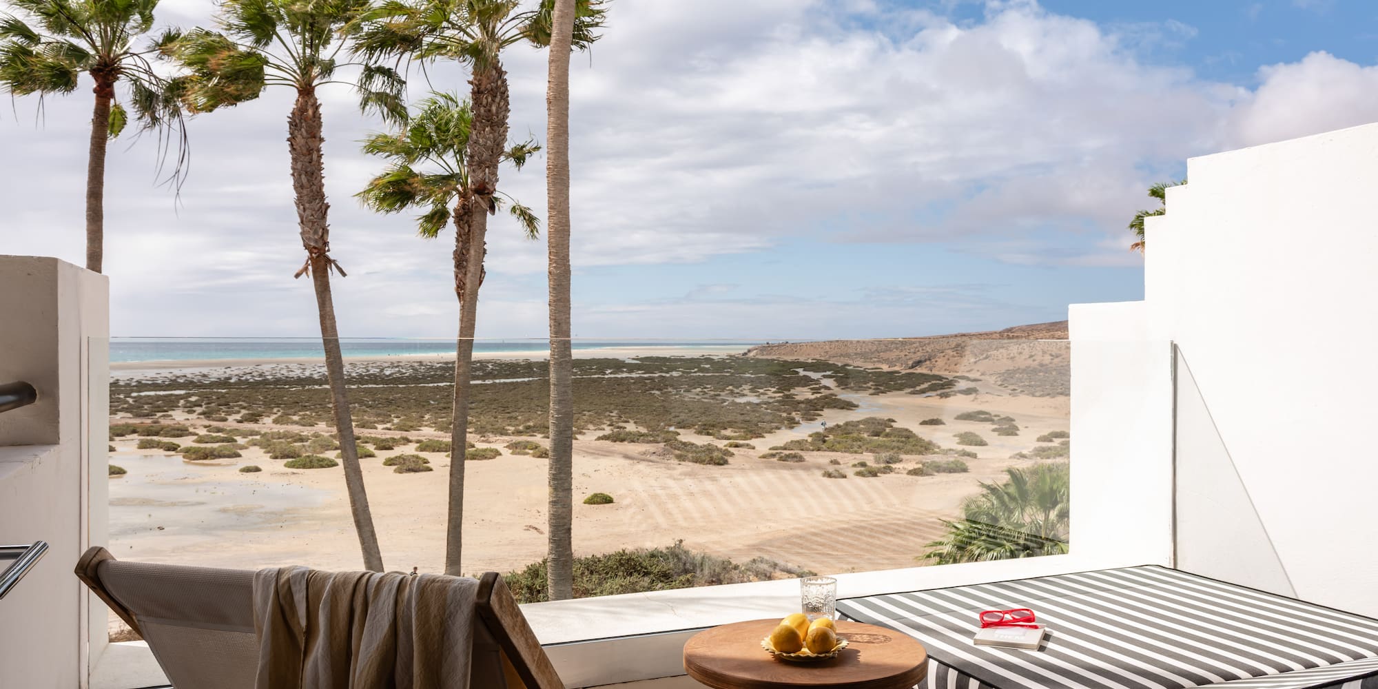 Sunny balcony overlooking sandy beach, ocean, palm trees, and lounge furniture.