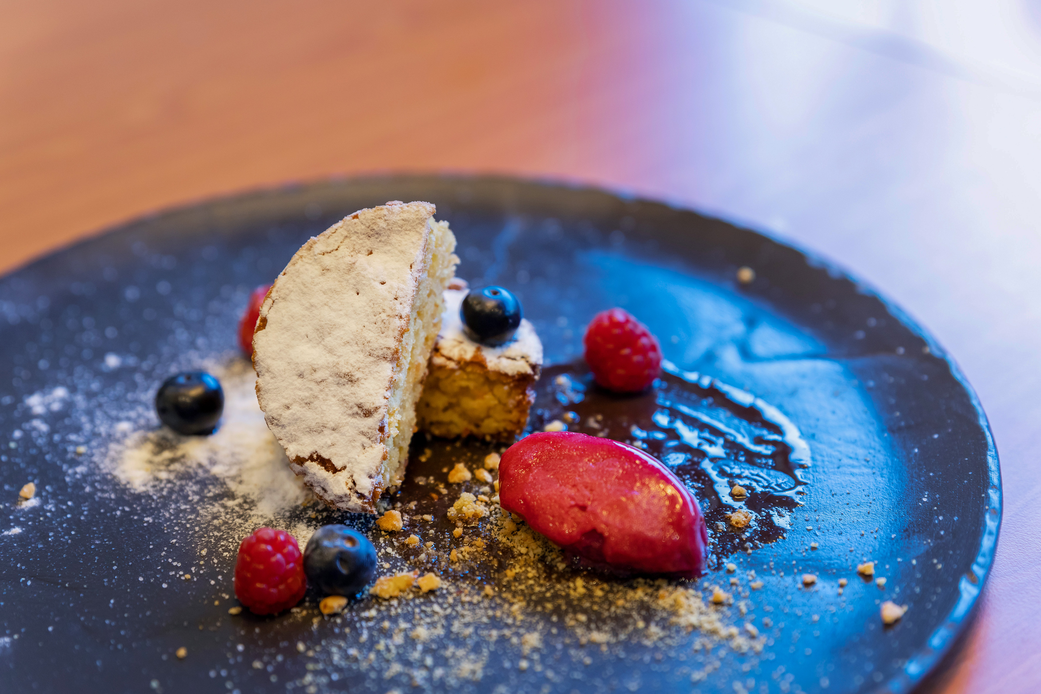 Powdered sugar cake, berries, and red sorbet dessert on dark plate.