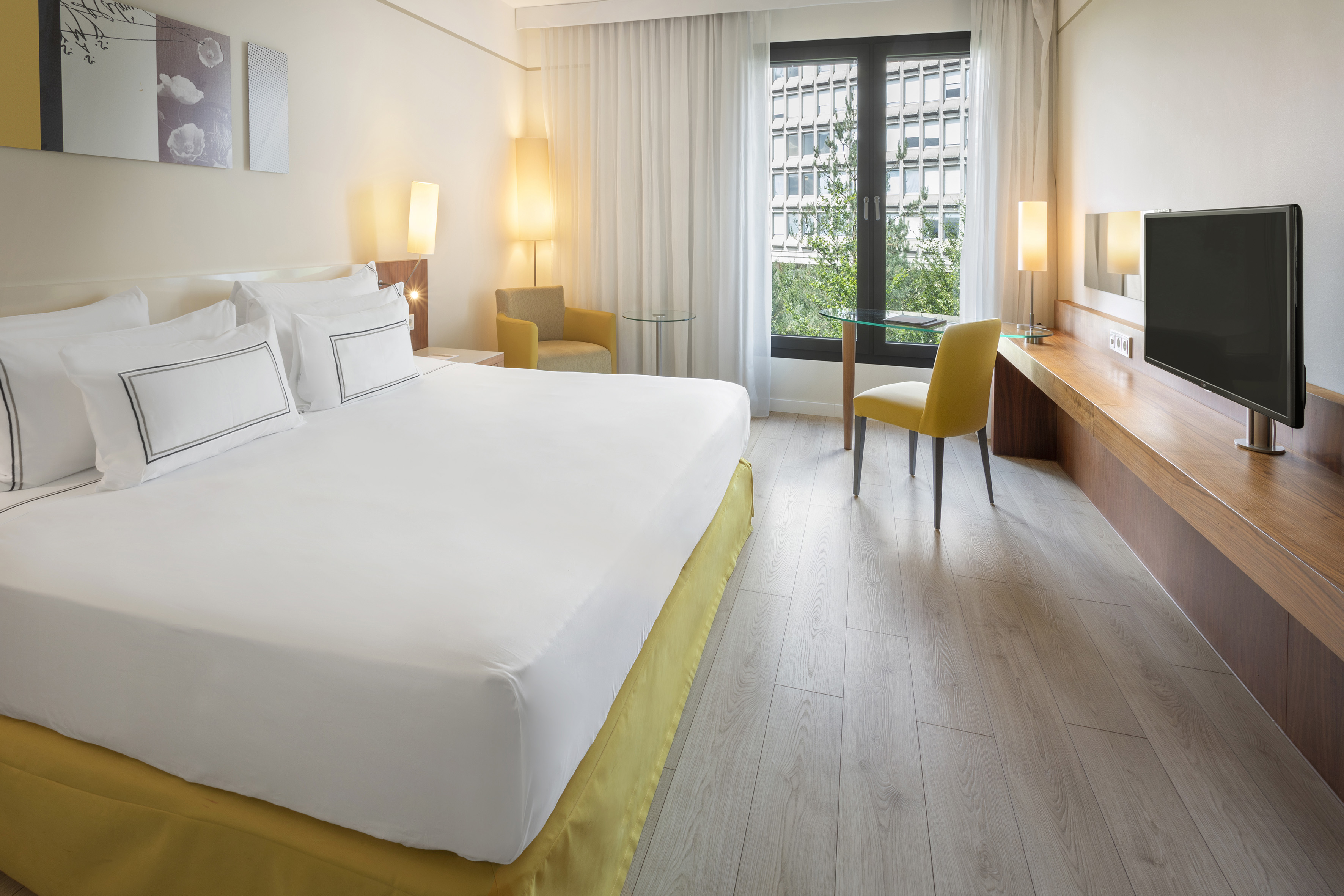 Modern hotel room with king bed, yellow armchair, desk, and city view.