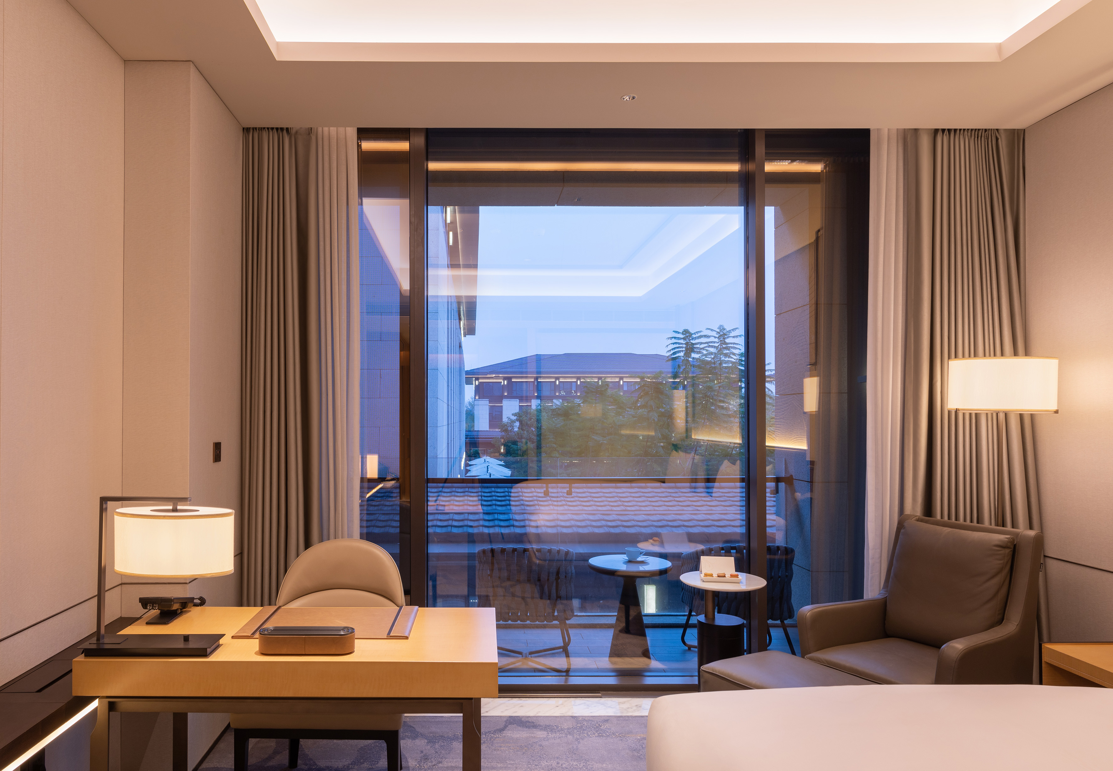 Modern luxury hotel room with desk, armchair, and balcony view.