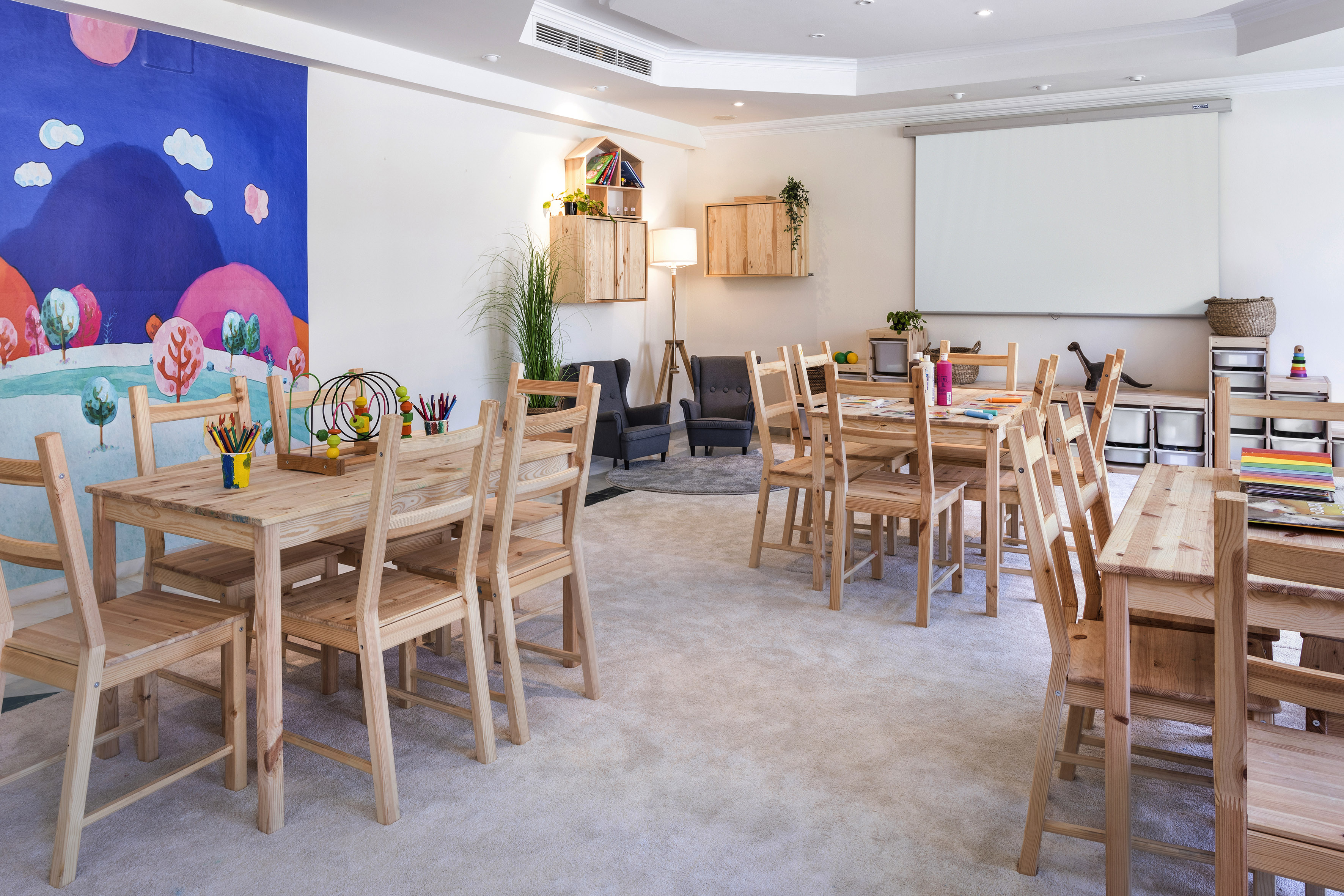 Bright children's play area with wooden tables, art supplies, colorful mural, screen.