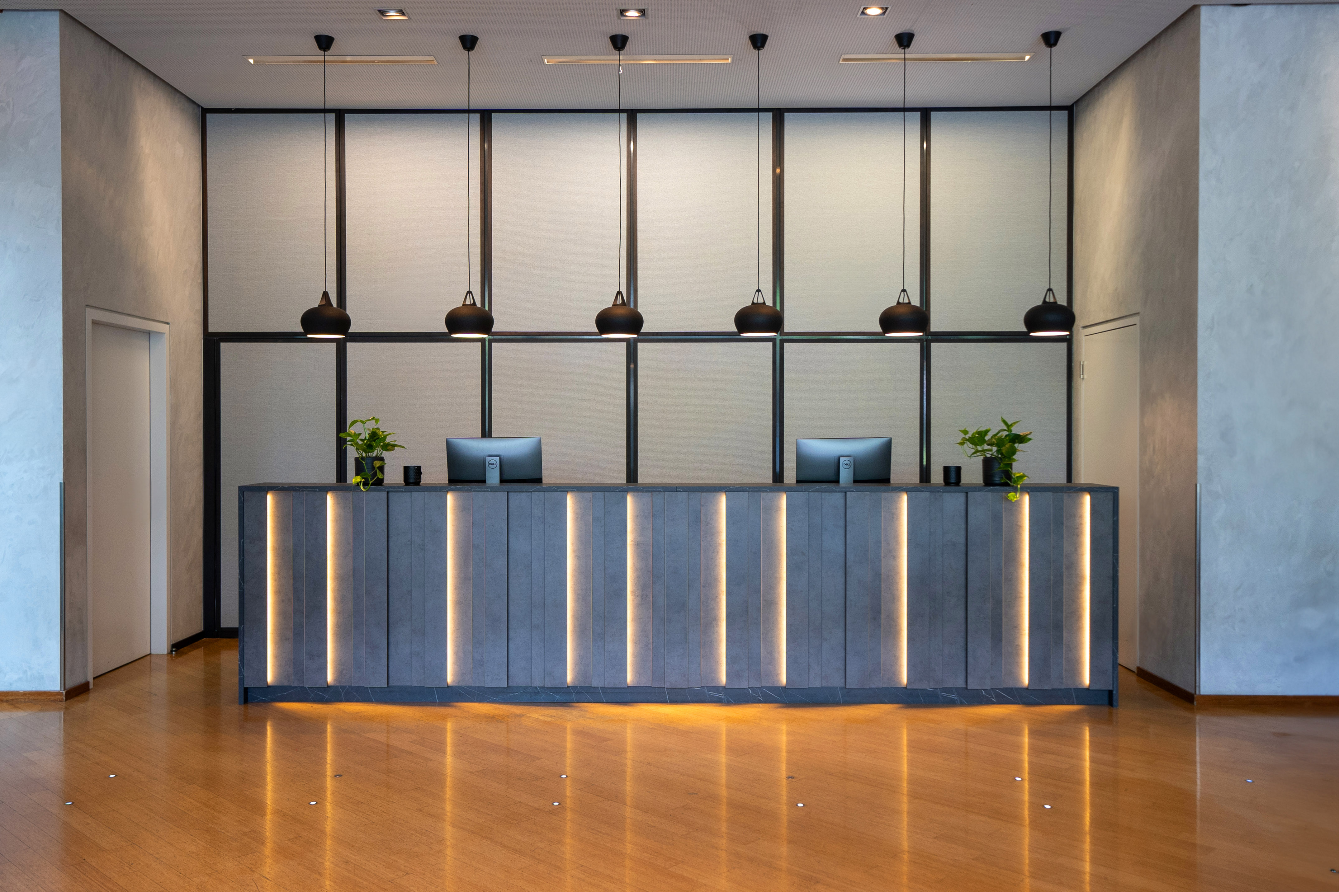 Modern hotel reception with illuminated grey desk, four monitors, pendant lights.