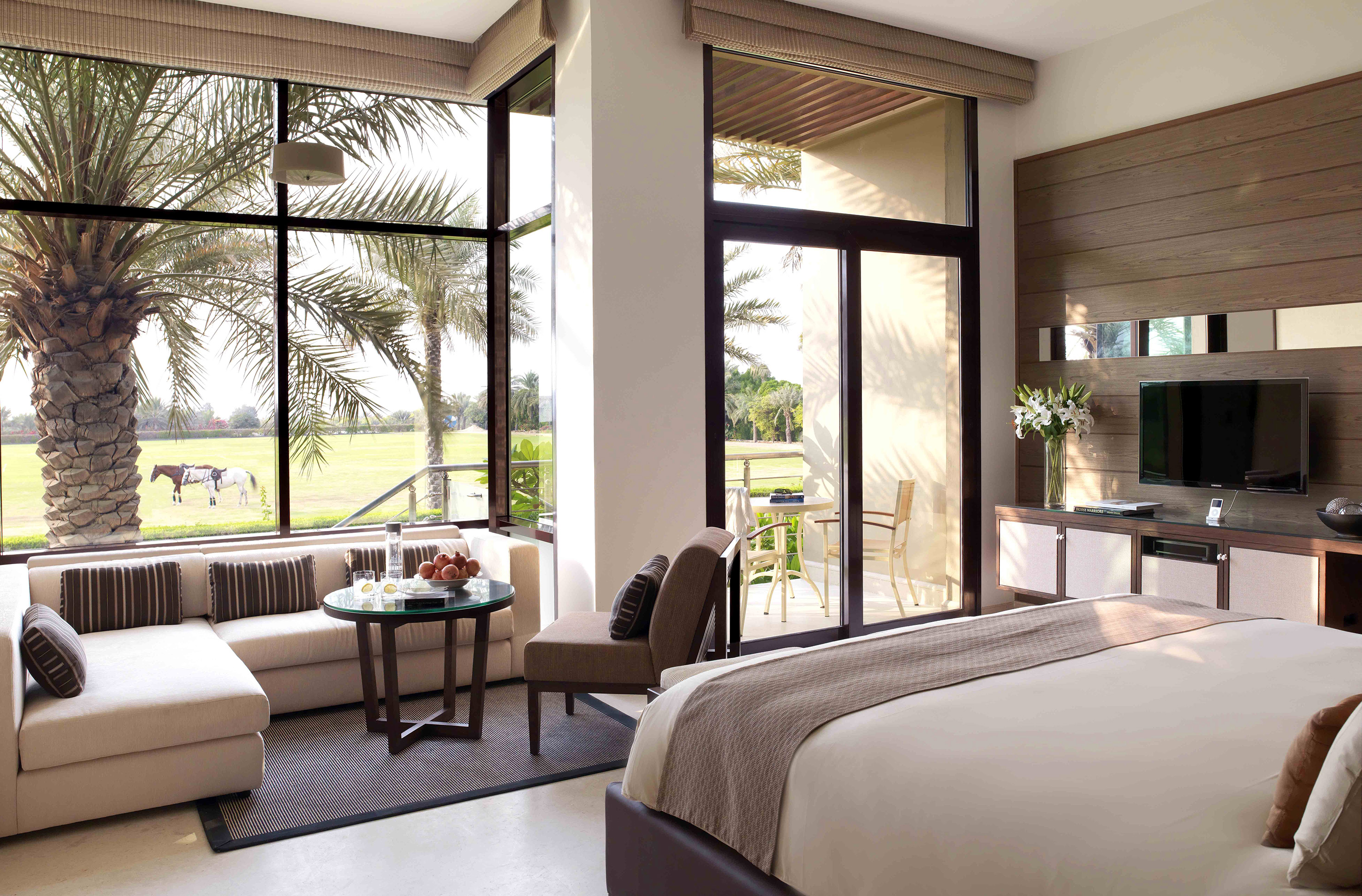 Elegant hotel room overlooking polo field with horses, palm trees, and balcony.