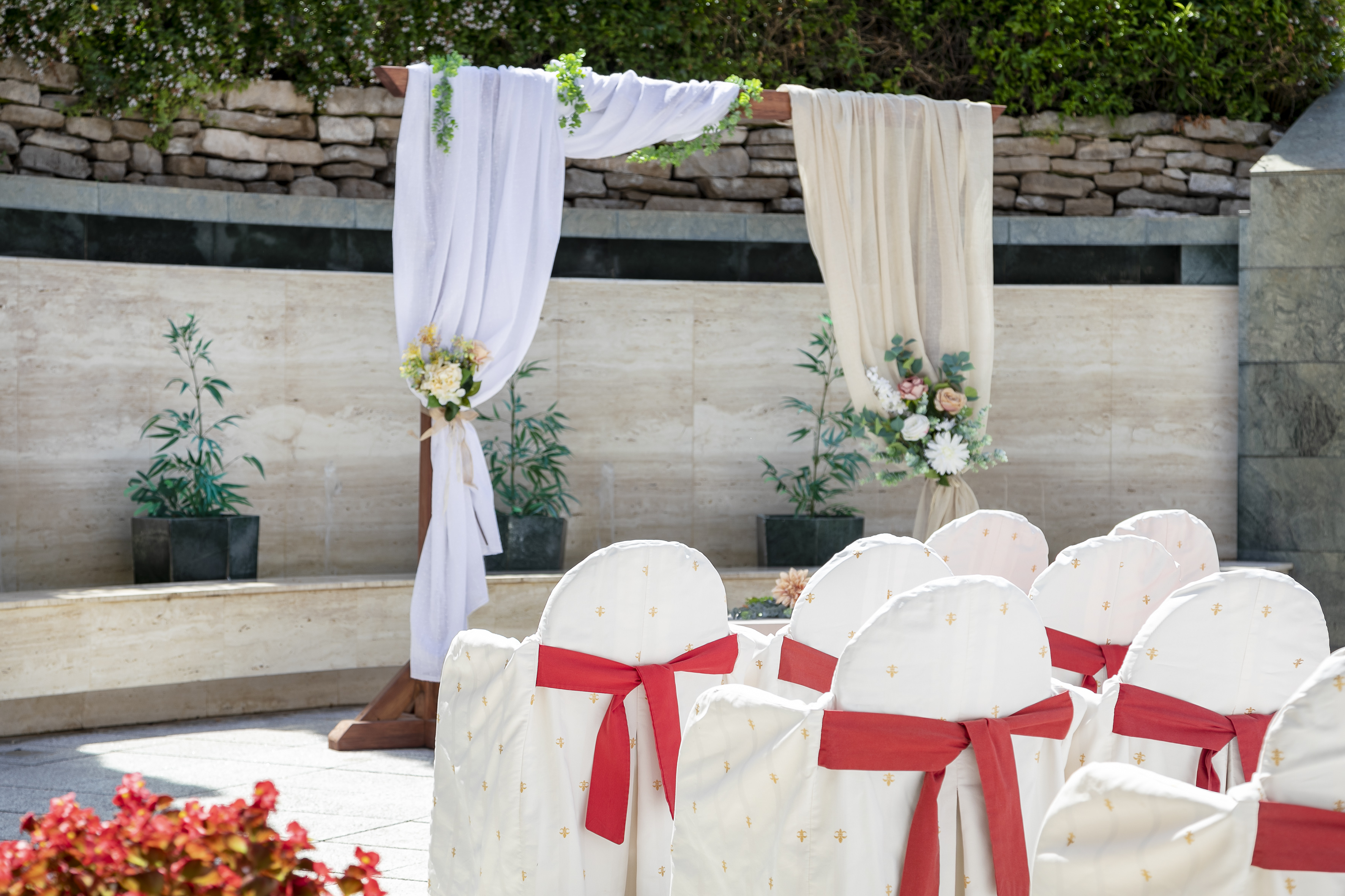 Outdoor wedding ceremony: draped arch with flowers, white chairs, red sashes.