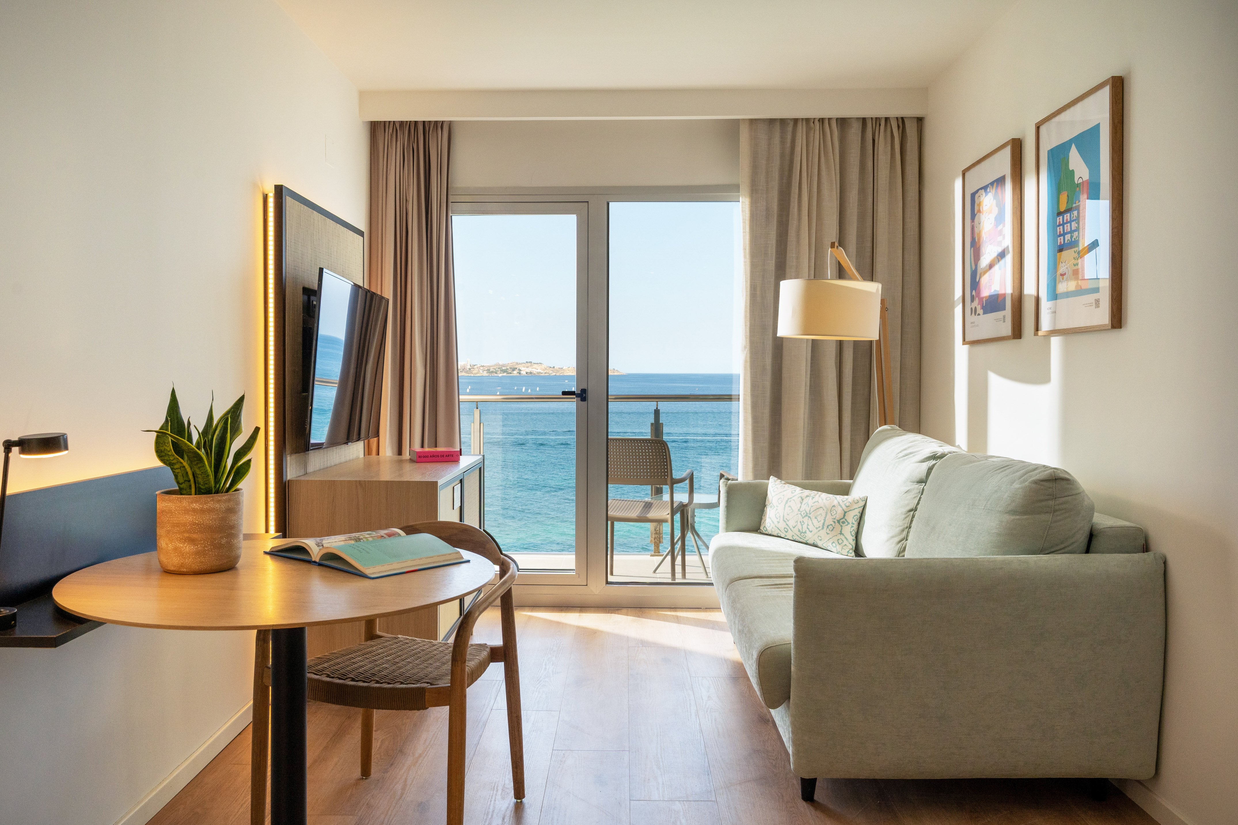 Modern hotel room with sea view balcony, sofa, TV, and workspace.