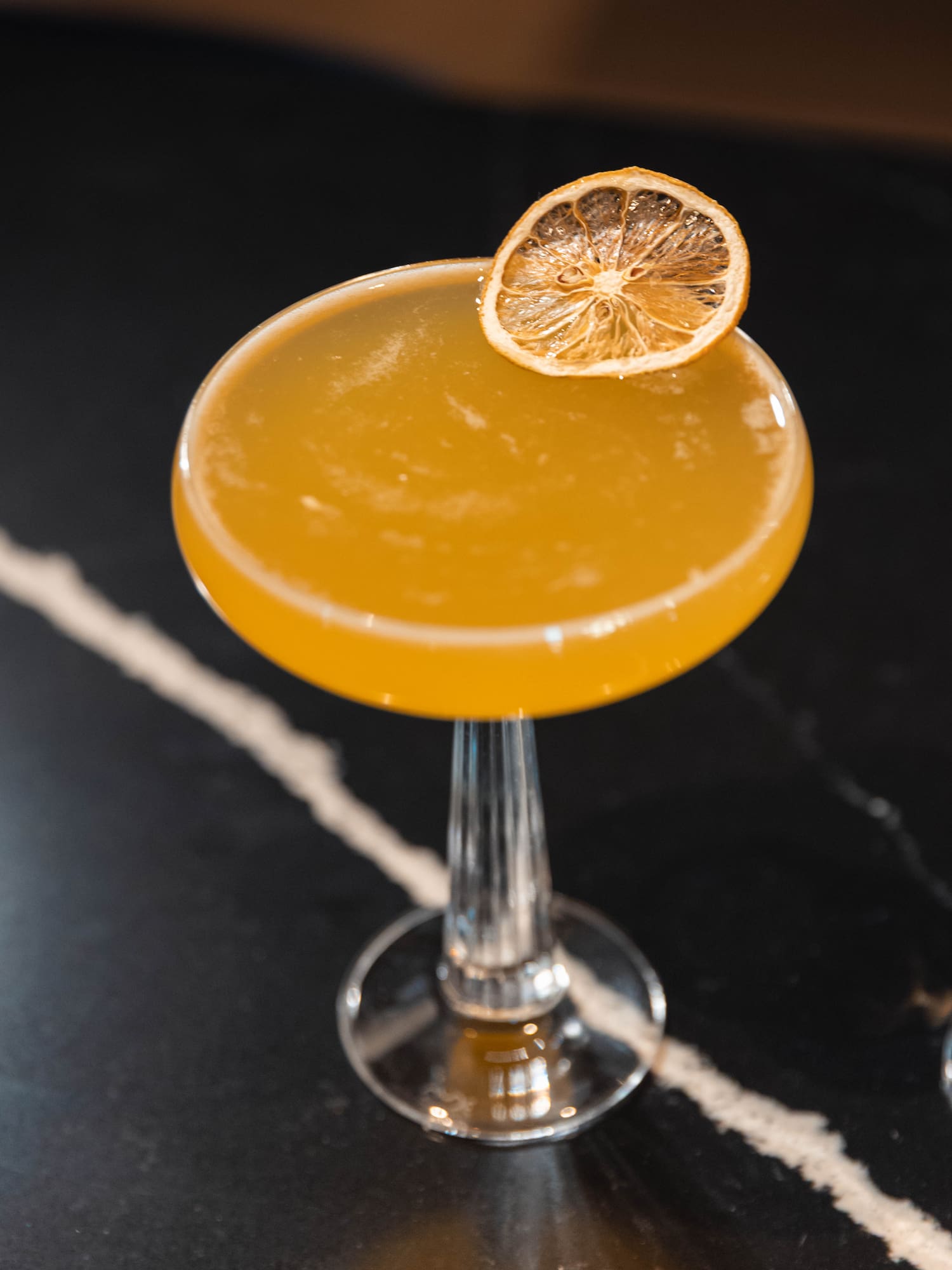 Yellow cocktail in coupe glass with dried orange slice on dark marble surface.