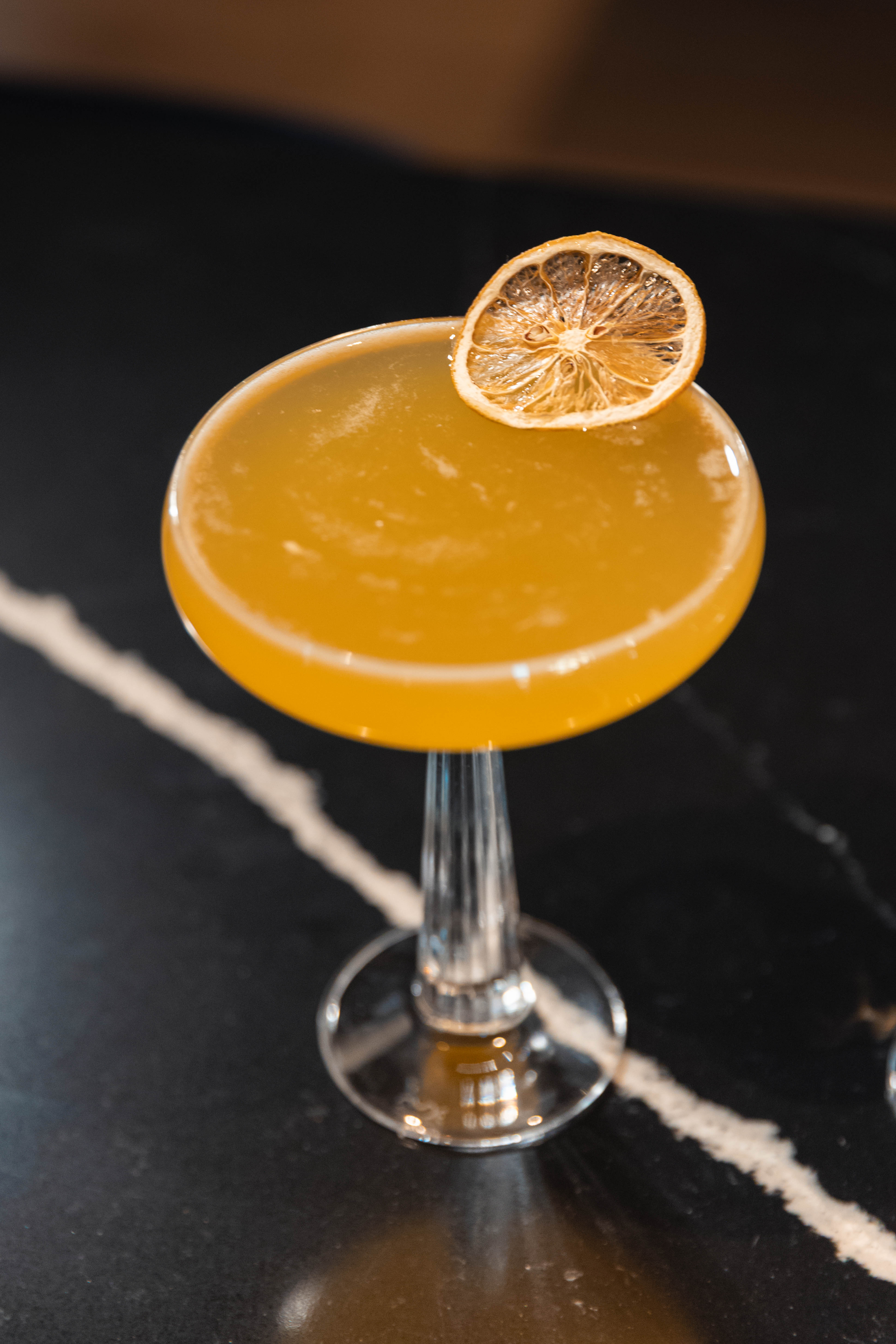 Yellow cocktail in coupe glass with dried orange slice on dark marble surface.