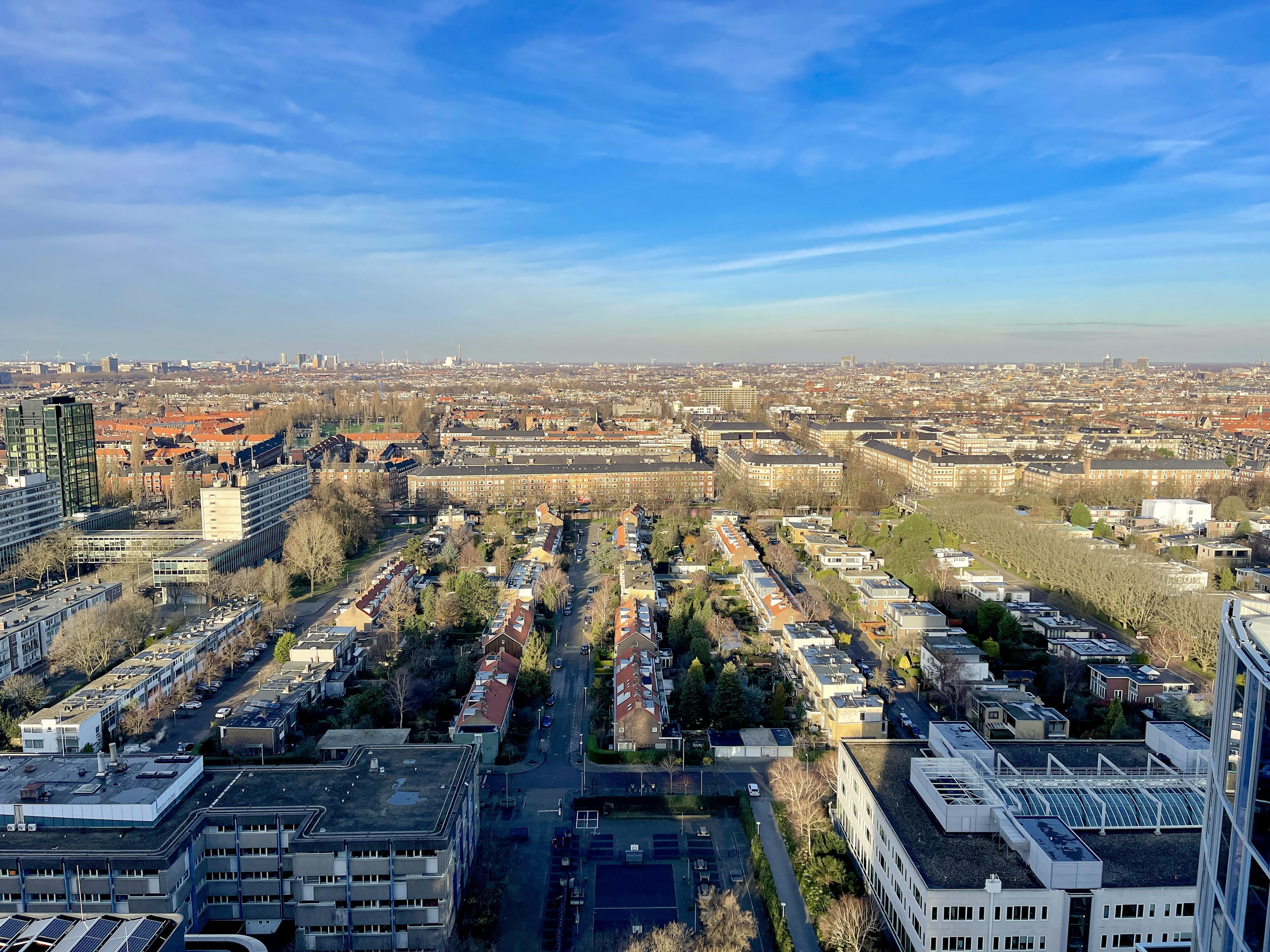 Aerial city view: residential buildings, streets, distant skyline, bright blue sky.