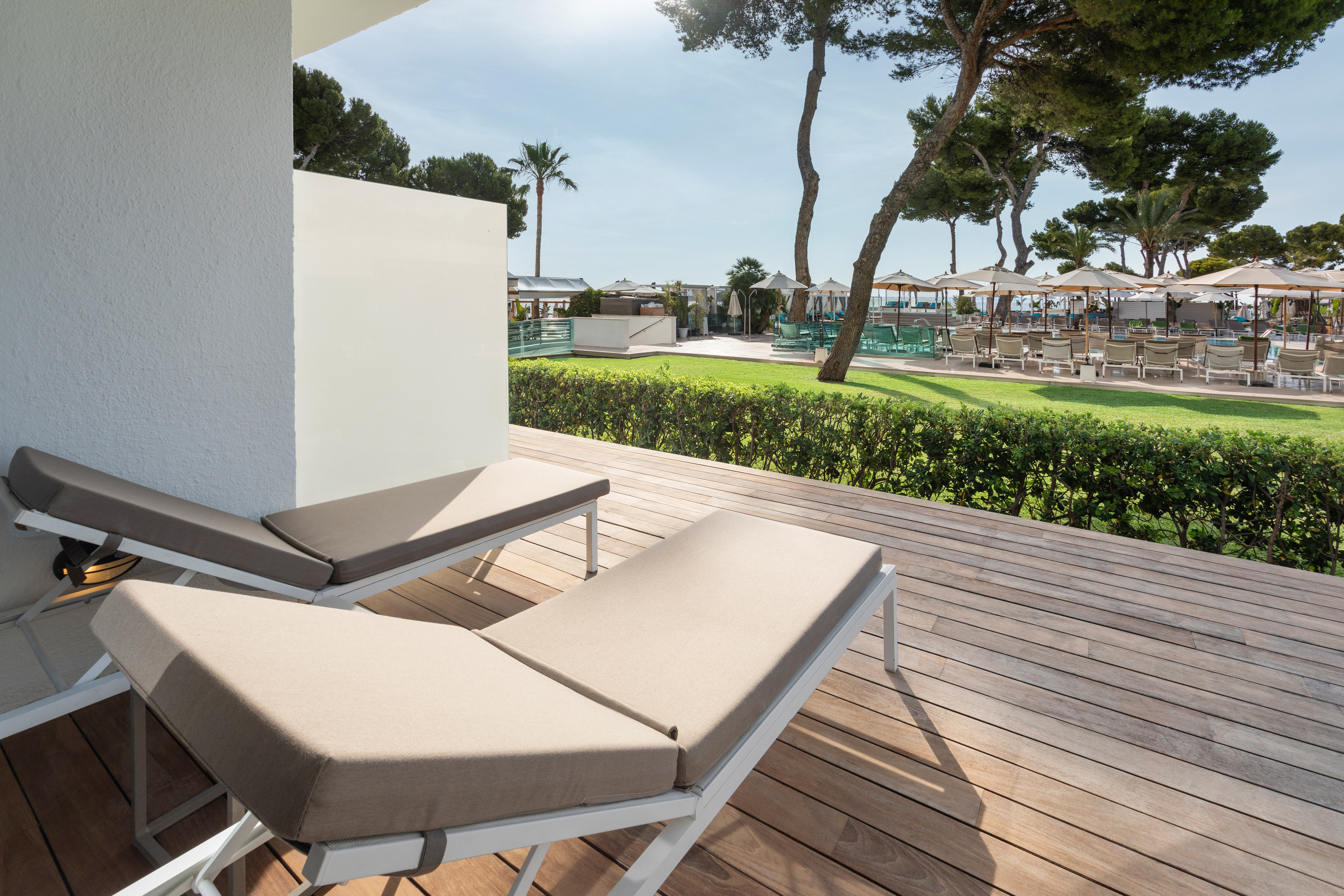 Two sun loungers on wooden deck overlooking sunny resort pool area with trees.