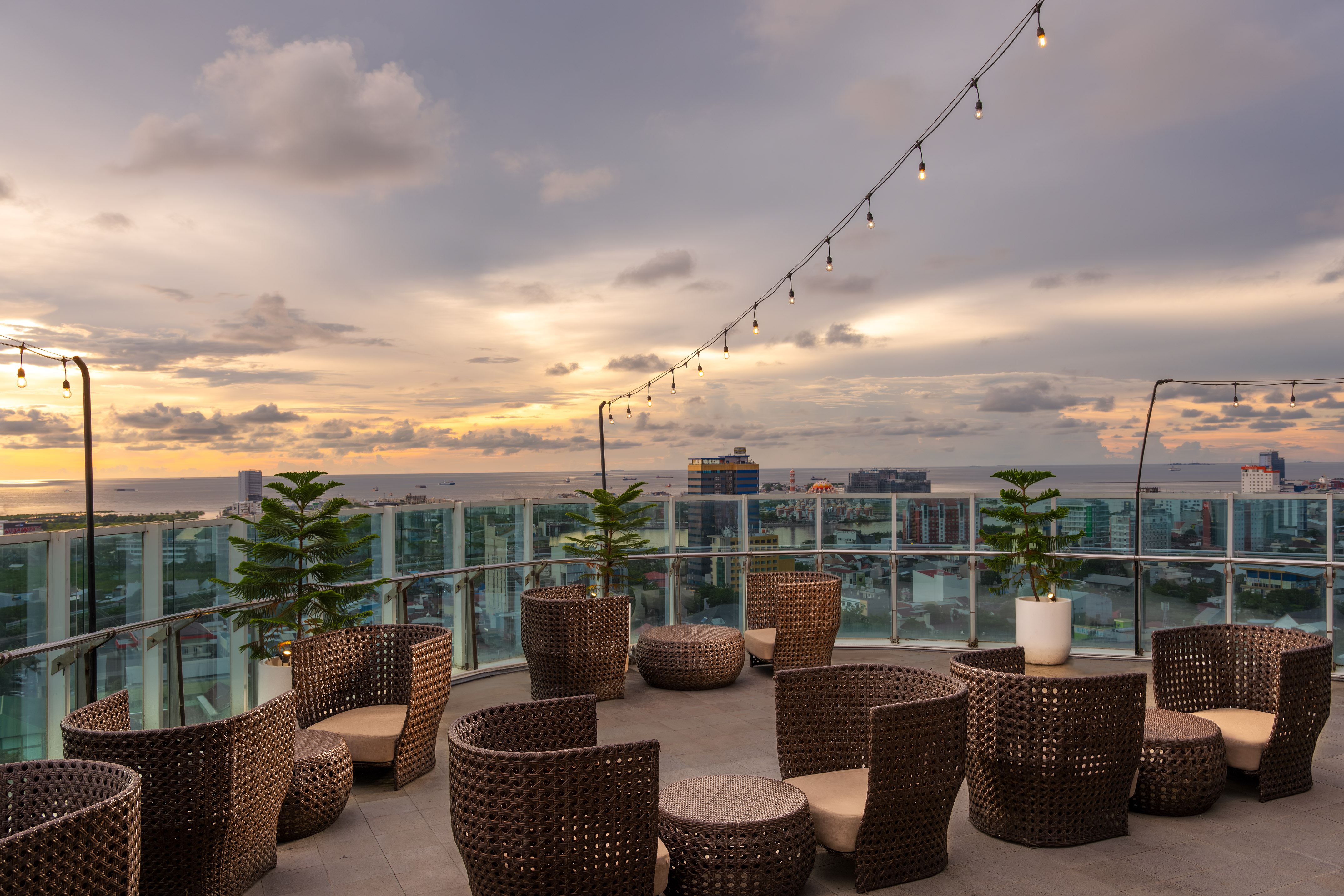 Rooftop lounge at sunset with woven chairs, string lights, city and ocean views.