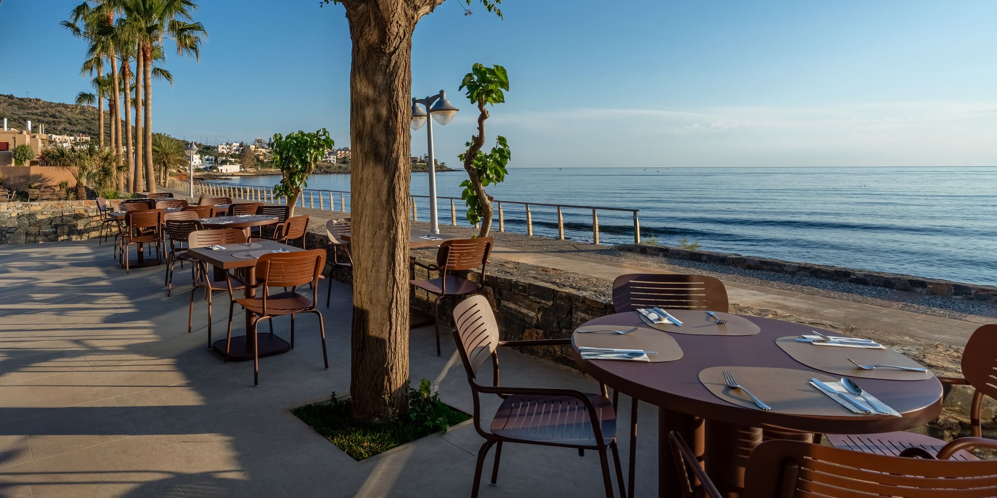 Coastal restaurant terrace with set tables, ocean view, and palm trees.