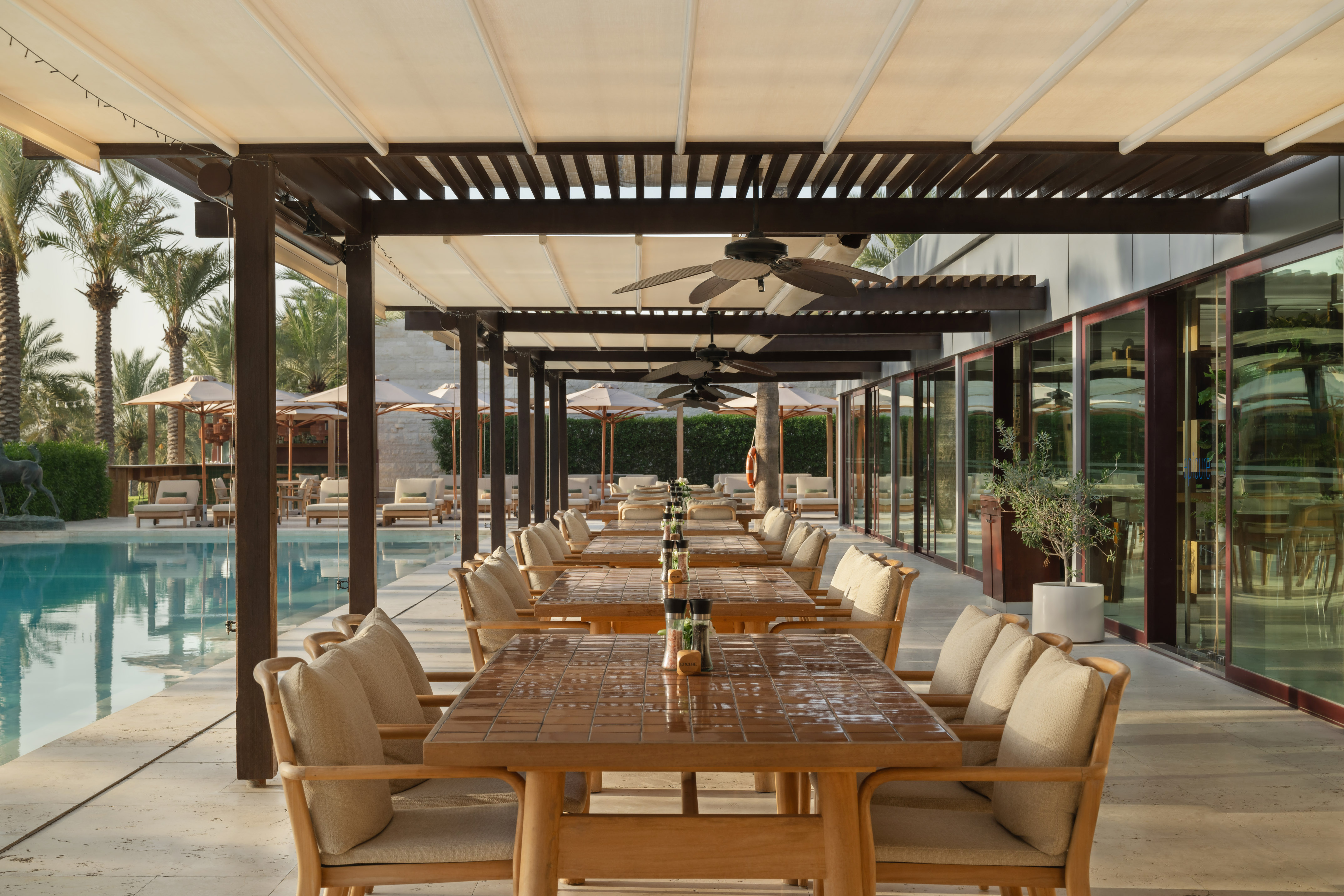 Poolside resort dining patio with covered tables, chairs, and palm trees.