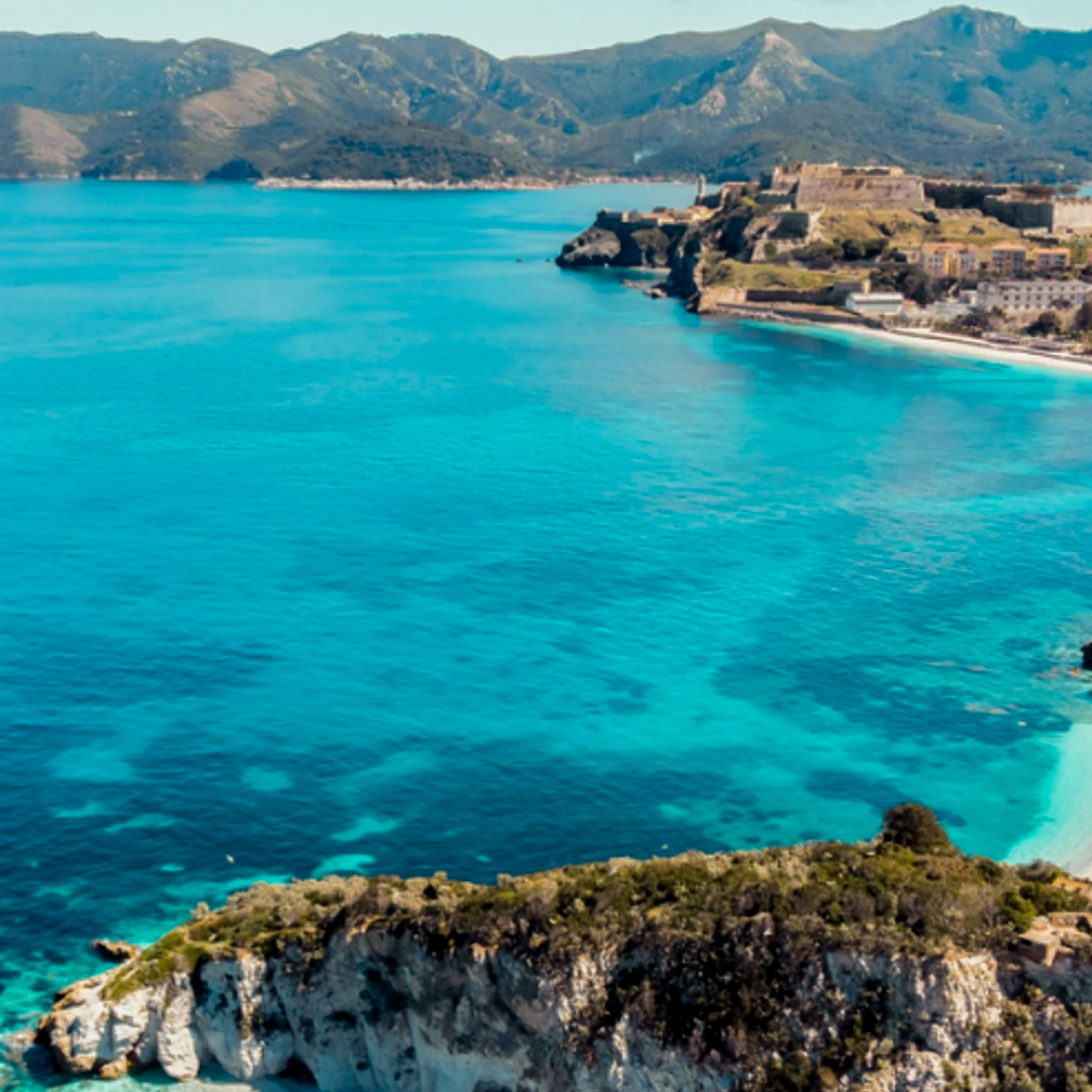 Turquoise sea with coastal town, ancient fortress, mountains, and rocky island.