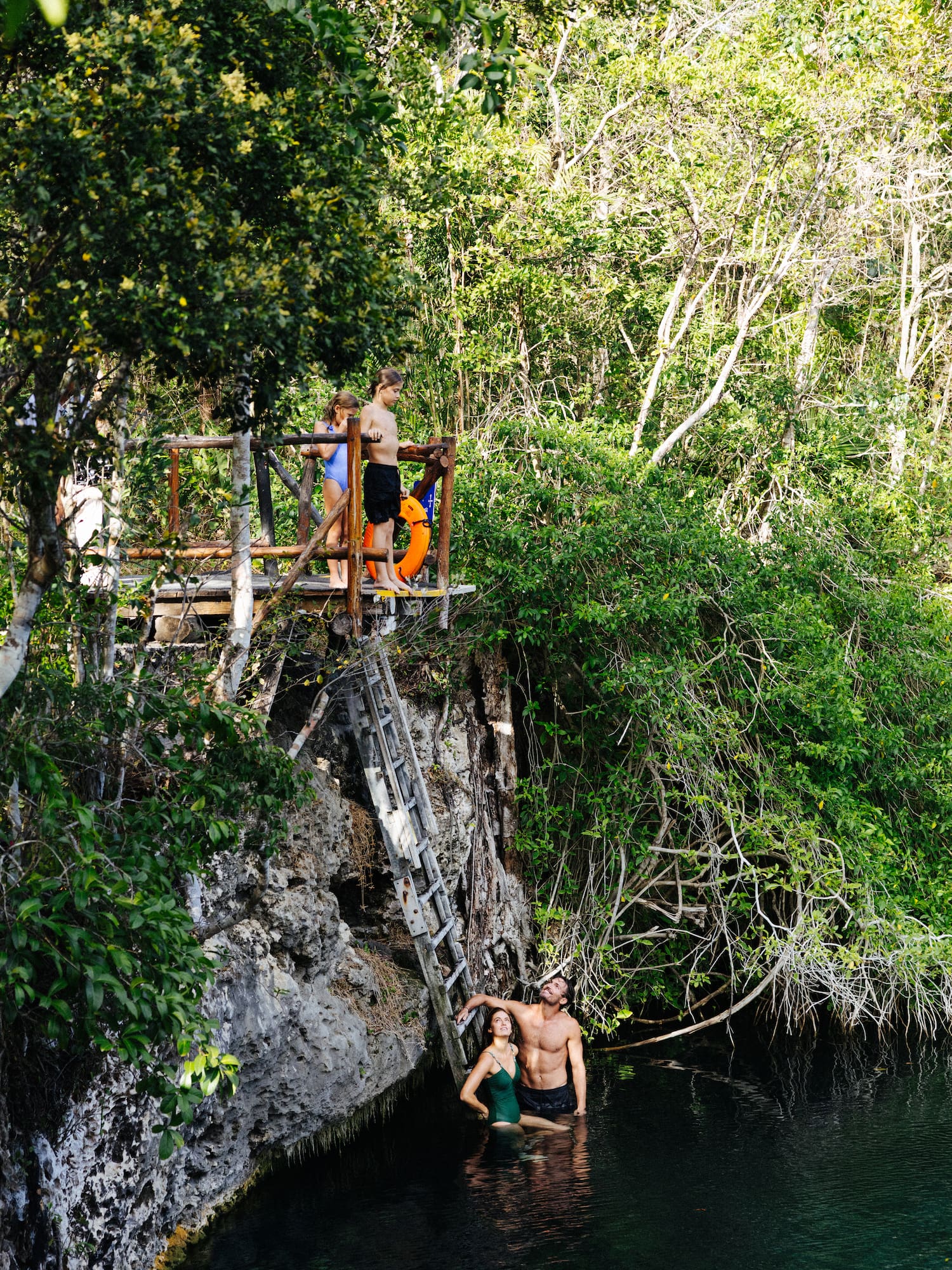 Family swimming in a jungle cenote, kids on a cliff jumping platform.