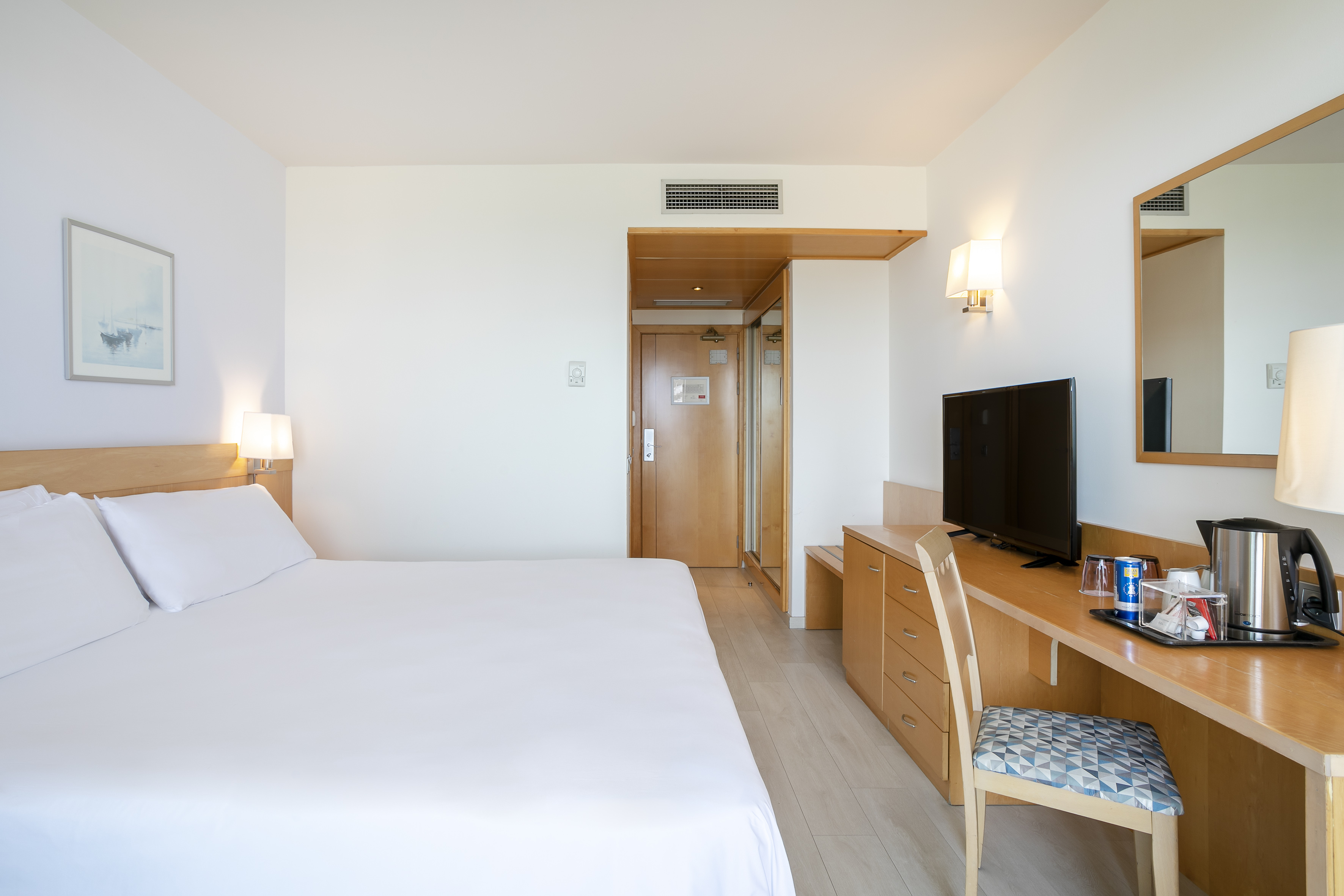 Modern hotel room with king bed, wooden desk, TV, and light wood floor.