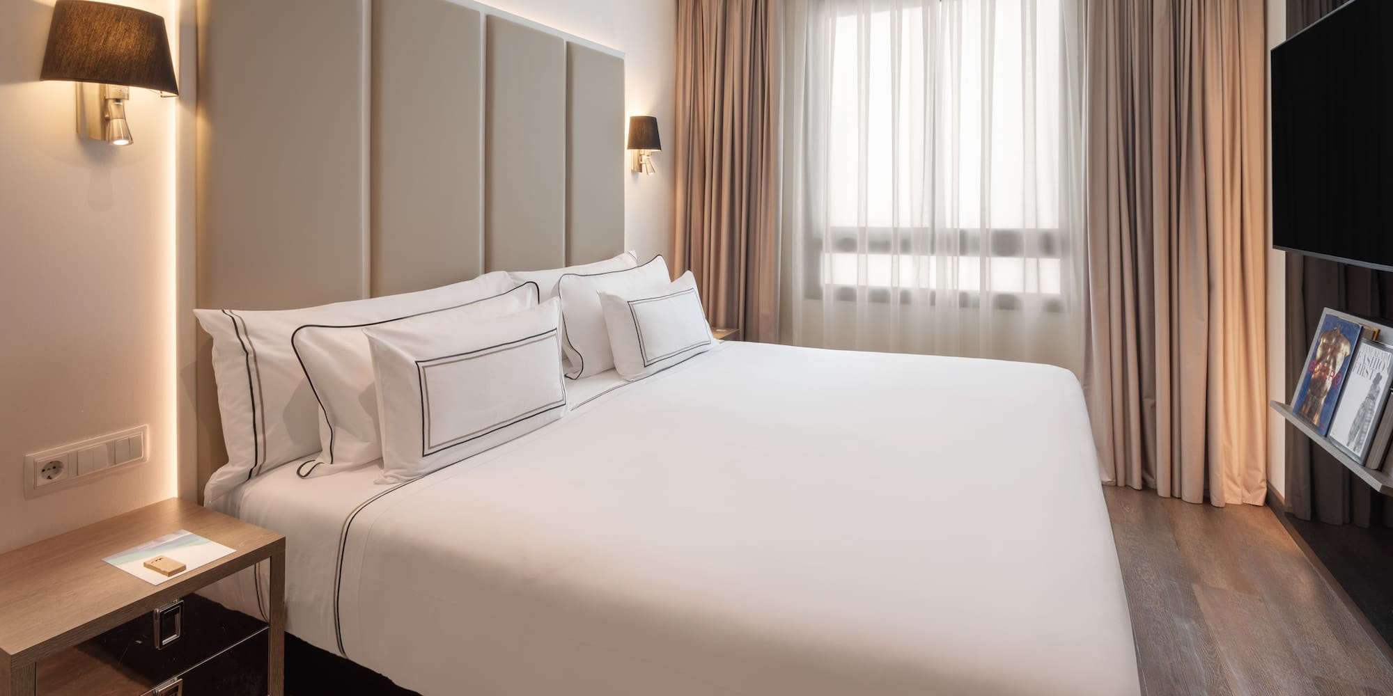 Modern hotel bedroom with king bed, white bedding, padded headboard, and window.
