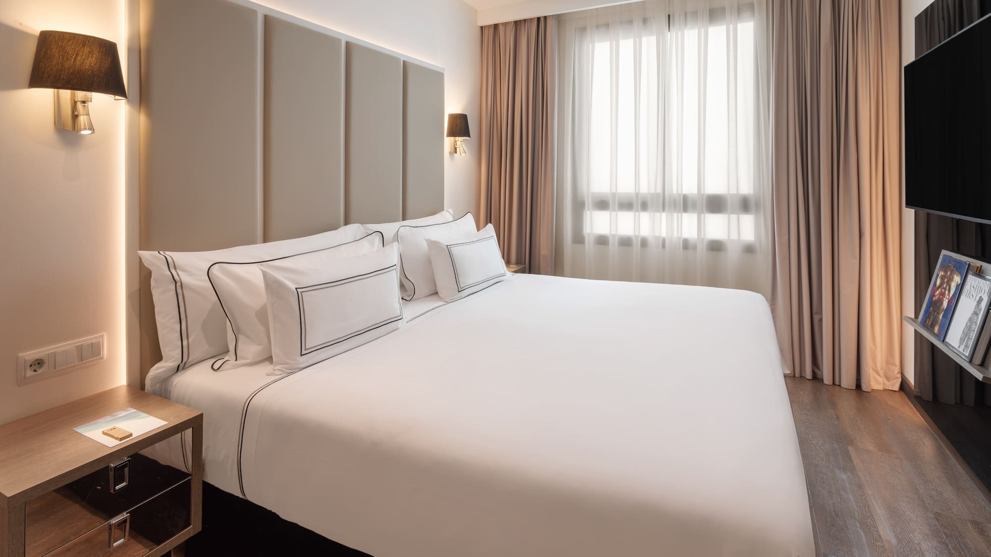 Modern hotel bedroom with king bed, white bedding, padded headboard, and window.