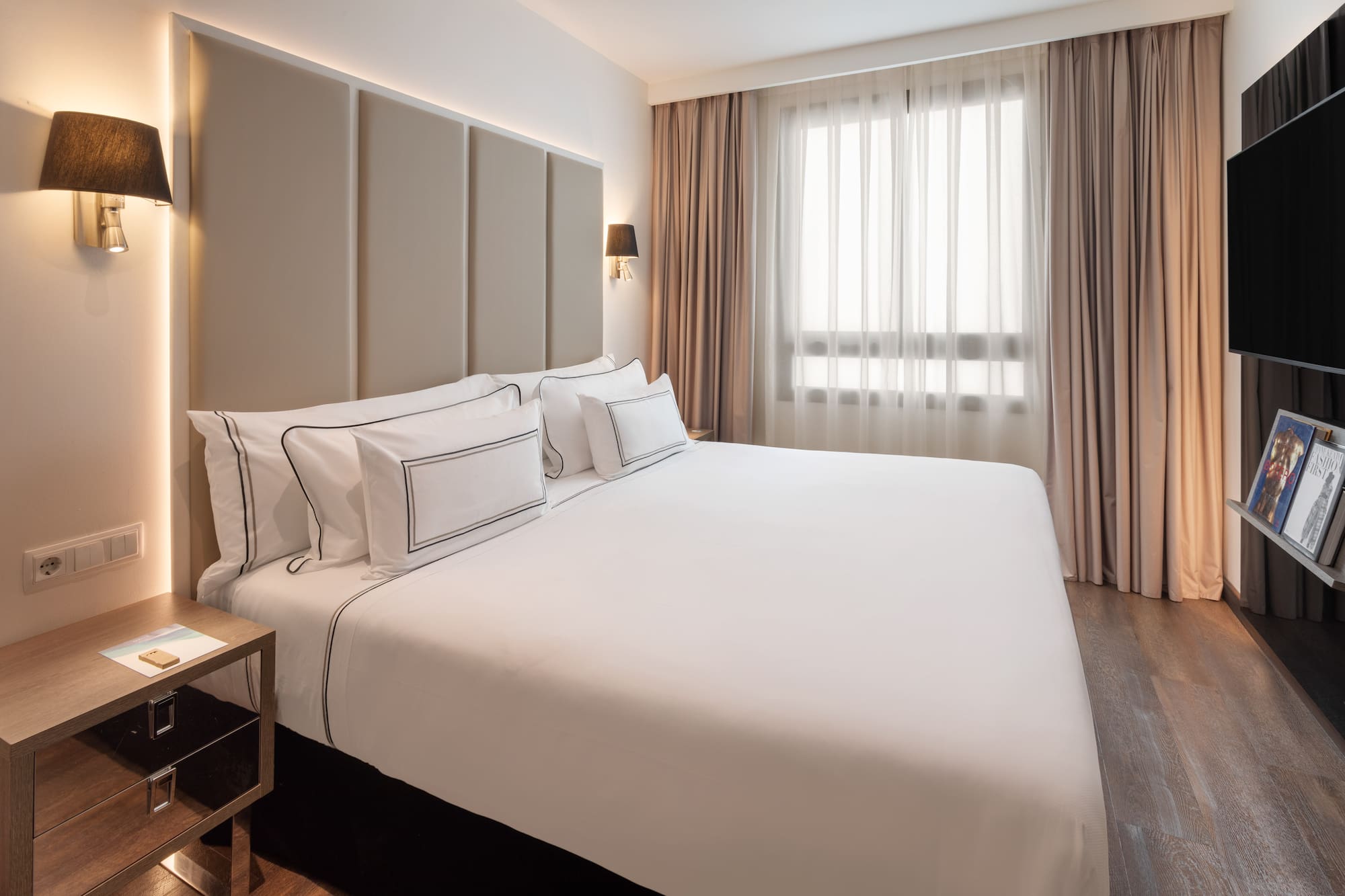 Modern hotel bedroom with king bed, white bedding, padded headboard, and window.