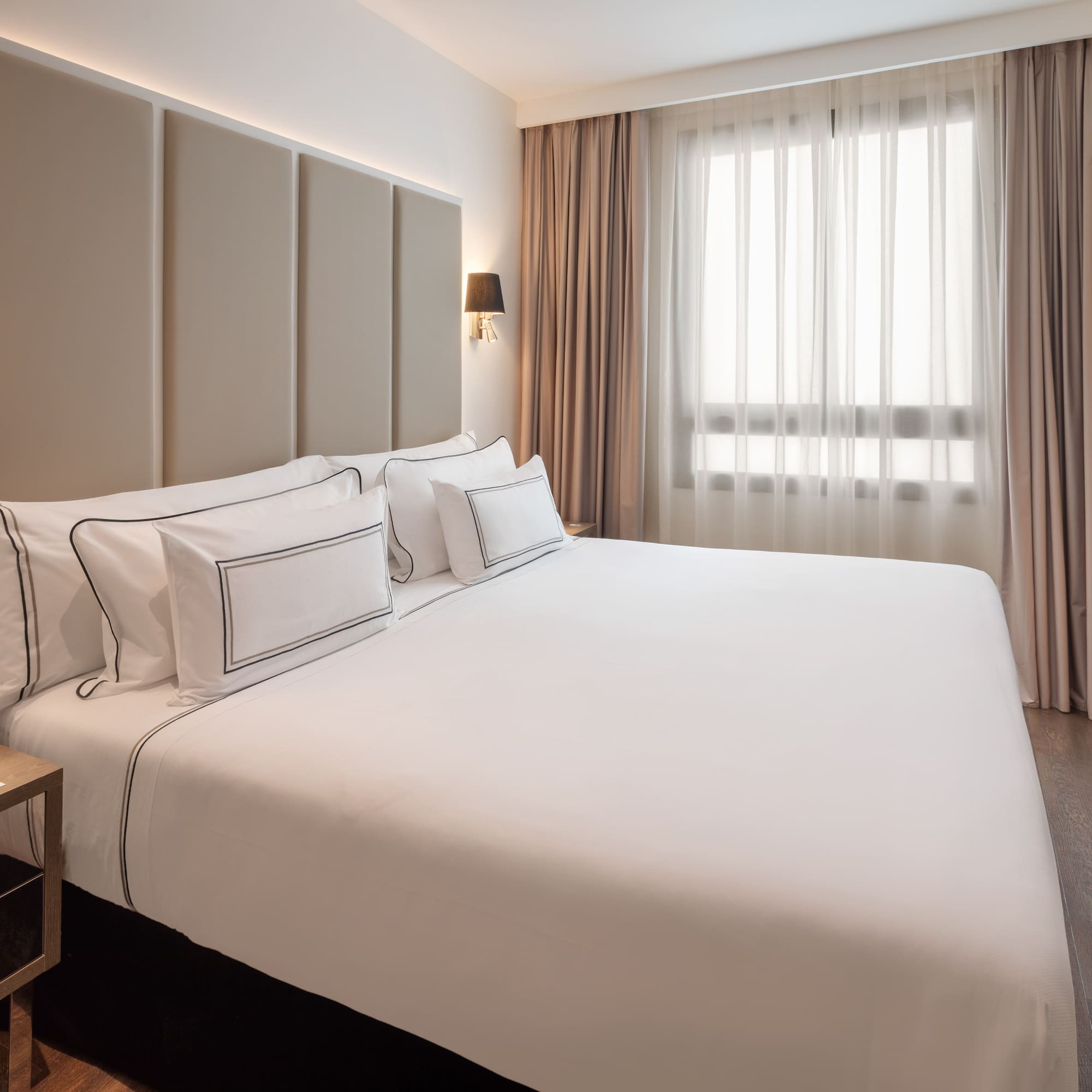 Modern hotel bedroom with king bed, white bedding, padded headboard, and window.