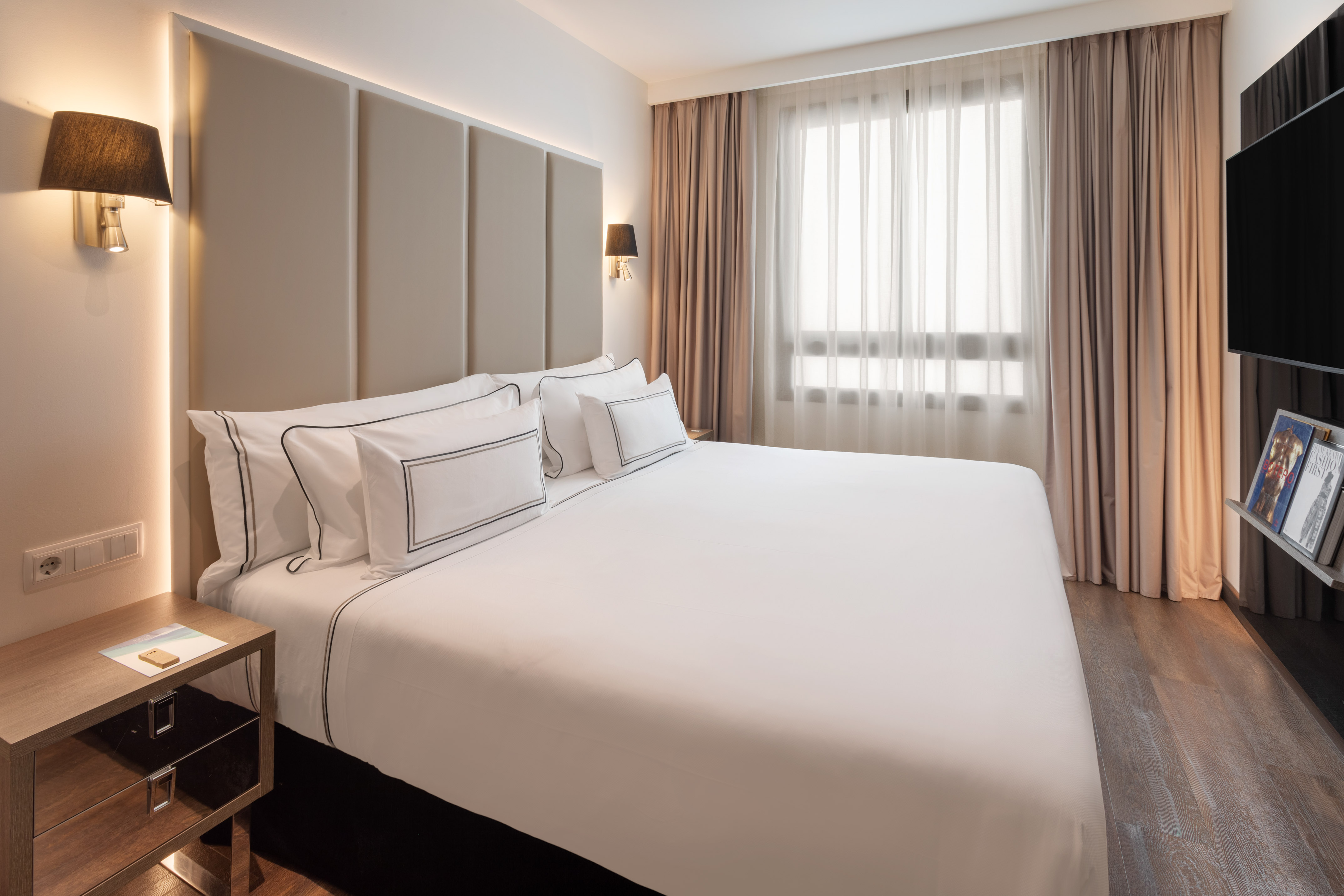 Modern hotel bedroom with king bed, white bedding, padded headboard, and window.