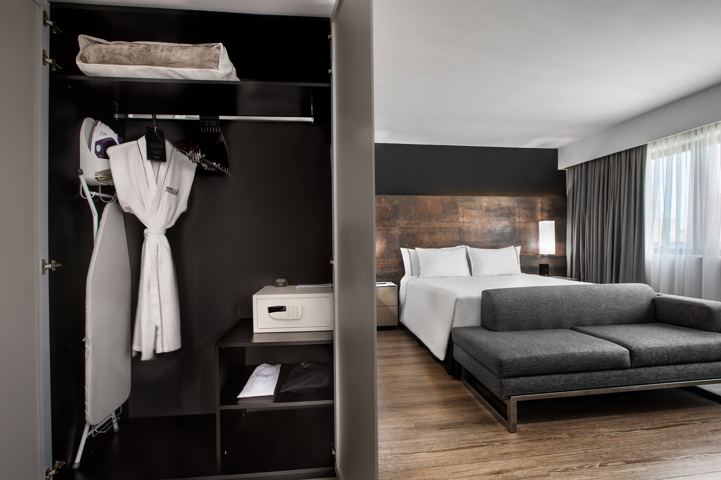 Modern hotel room with open wardrobe, iron, bathrobe, safe, bed, and sofa.