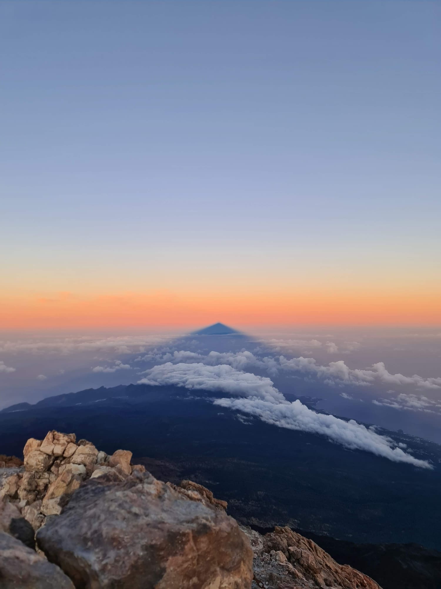 Teide volcano shadow over clouds at sunrise from rocky summit.