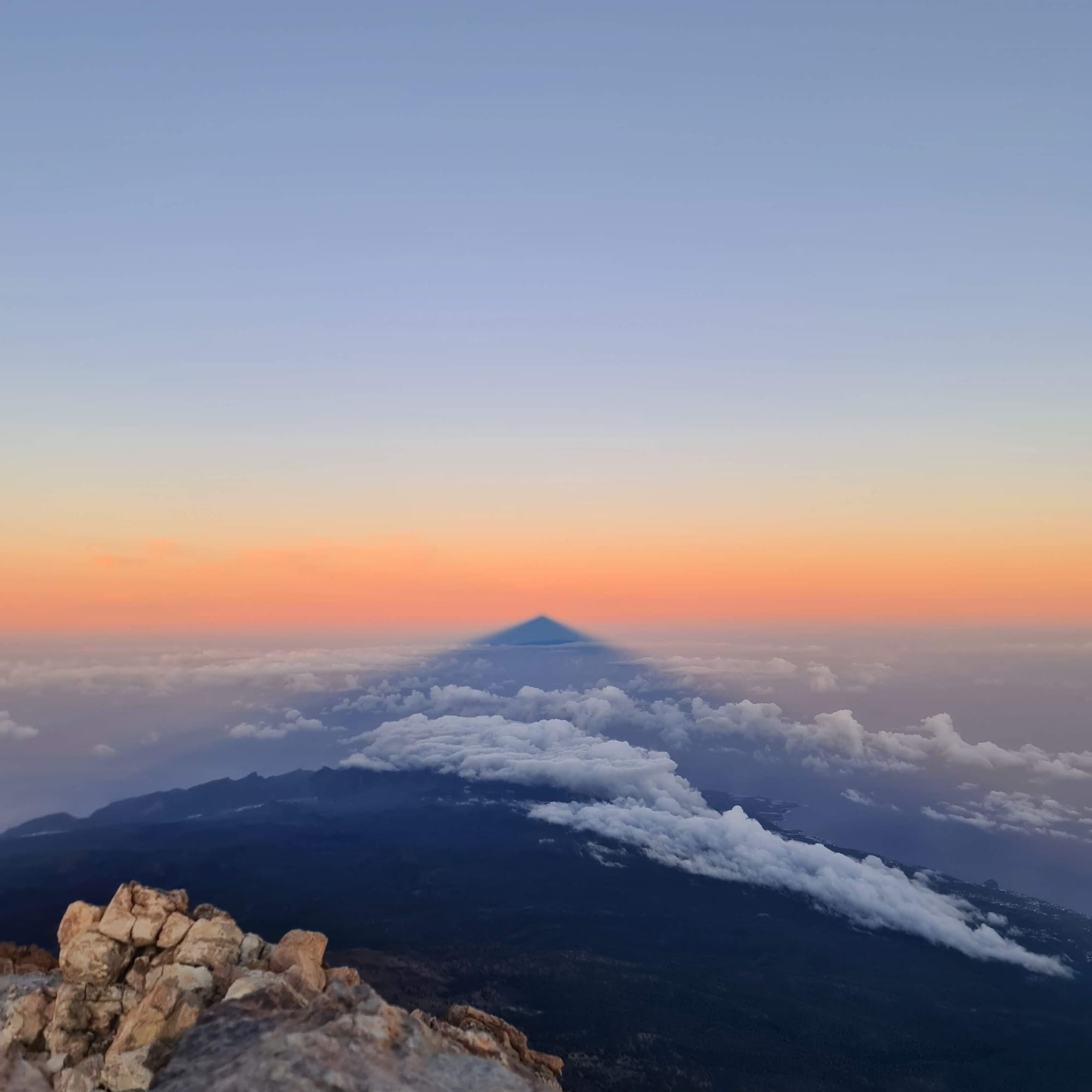 Teide volcano shadow over clouds at sunrise from rocky summit.