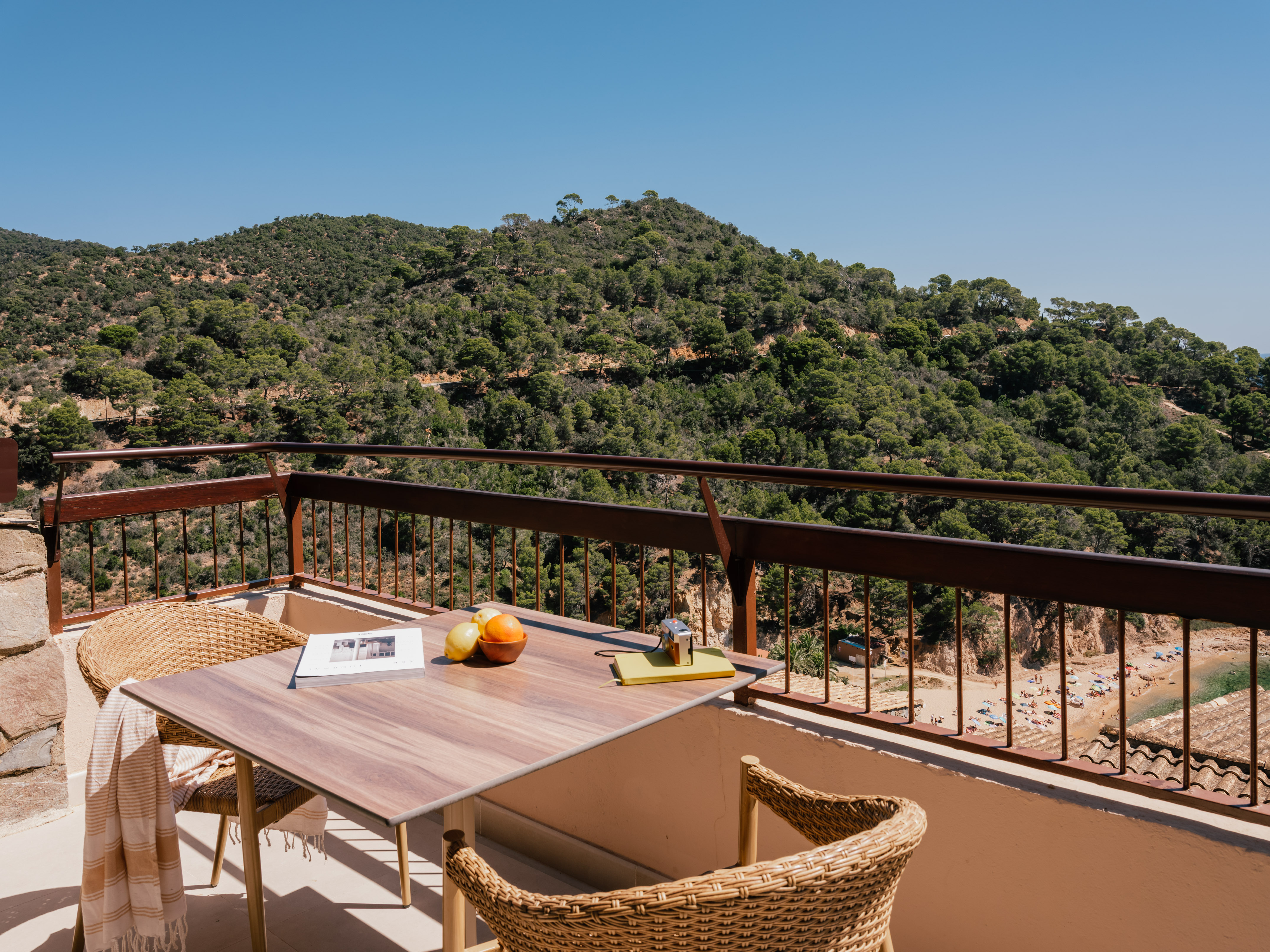 Balcony view of sunny Mediterranean beach, green mountains, table with fruit.