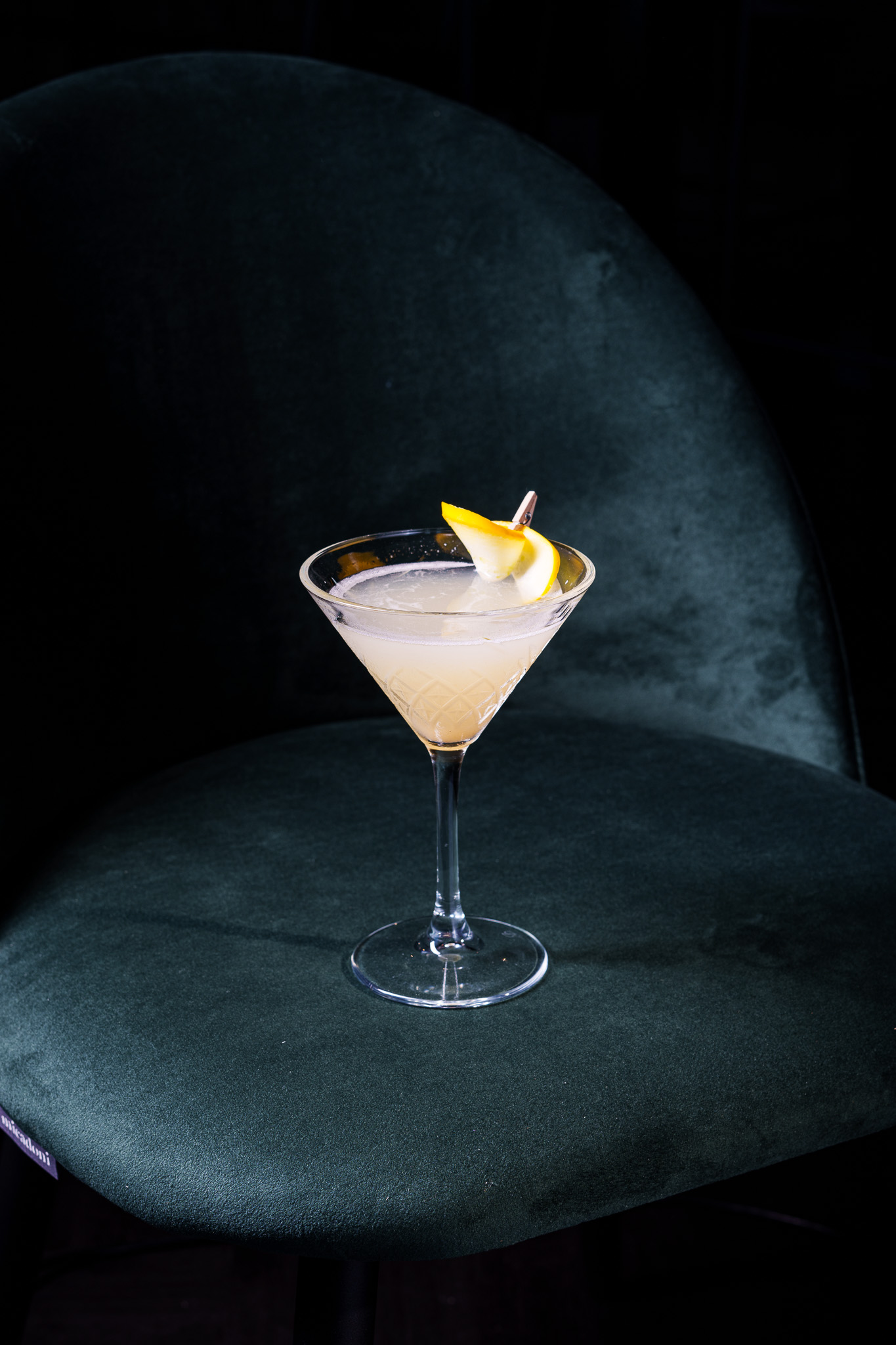 Martini cocktail with lemon garnish on dark green velvet chair.