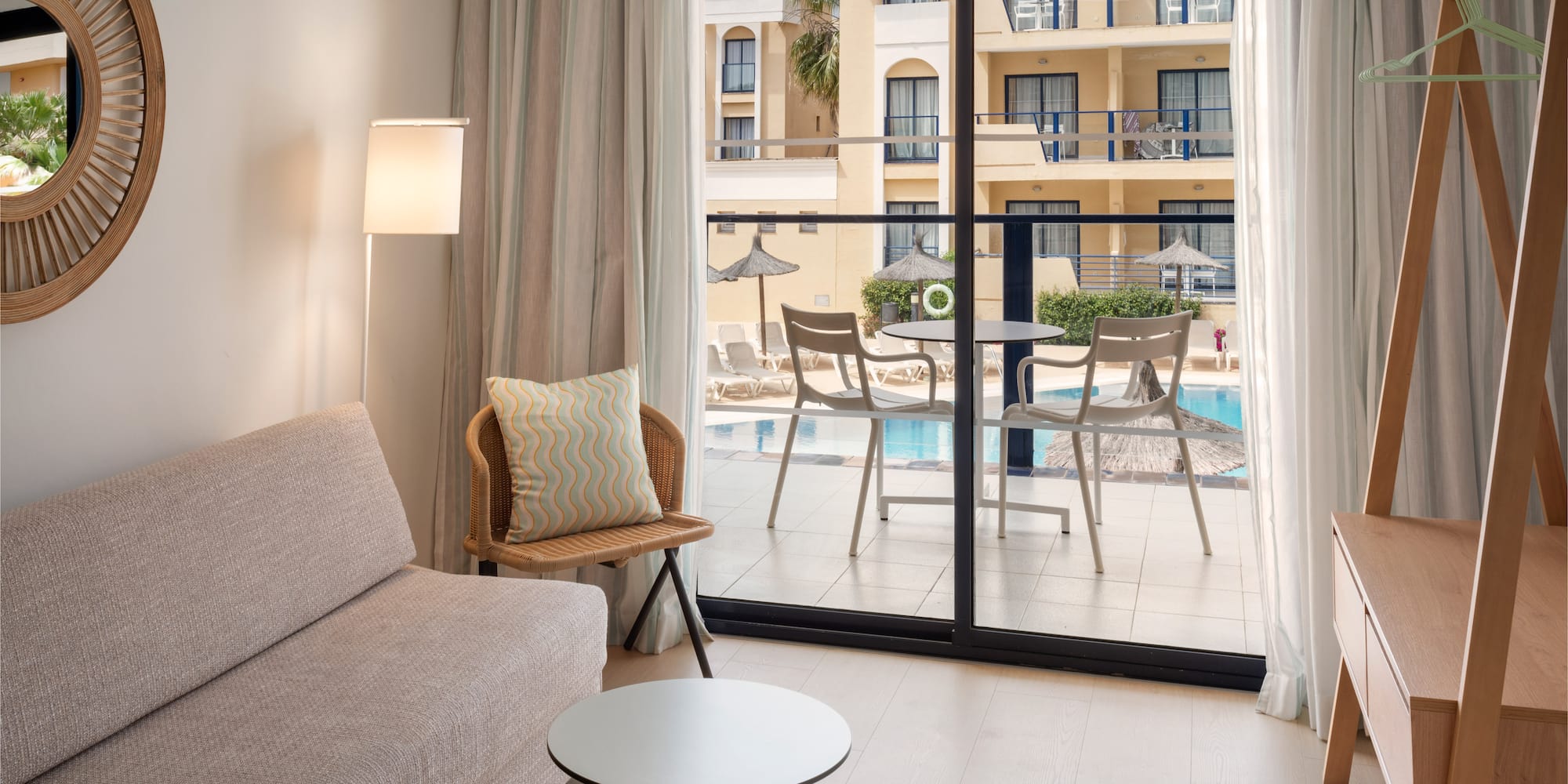 Modern hotel room with sofa, mirror, and balcony overlooking a resort pool.