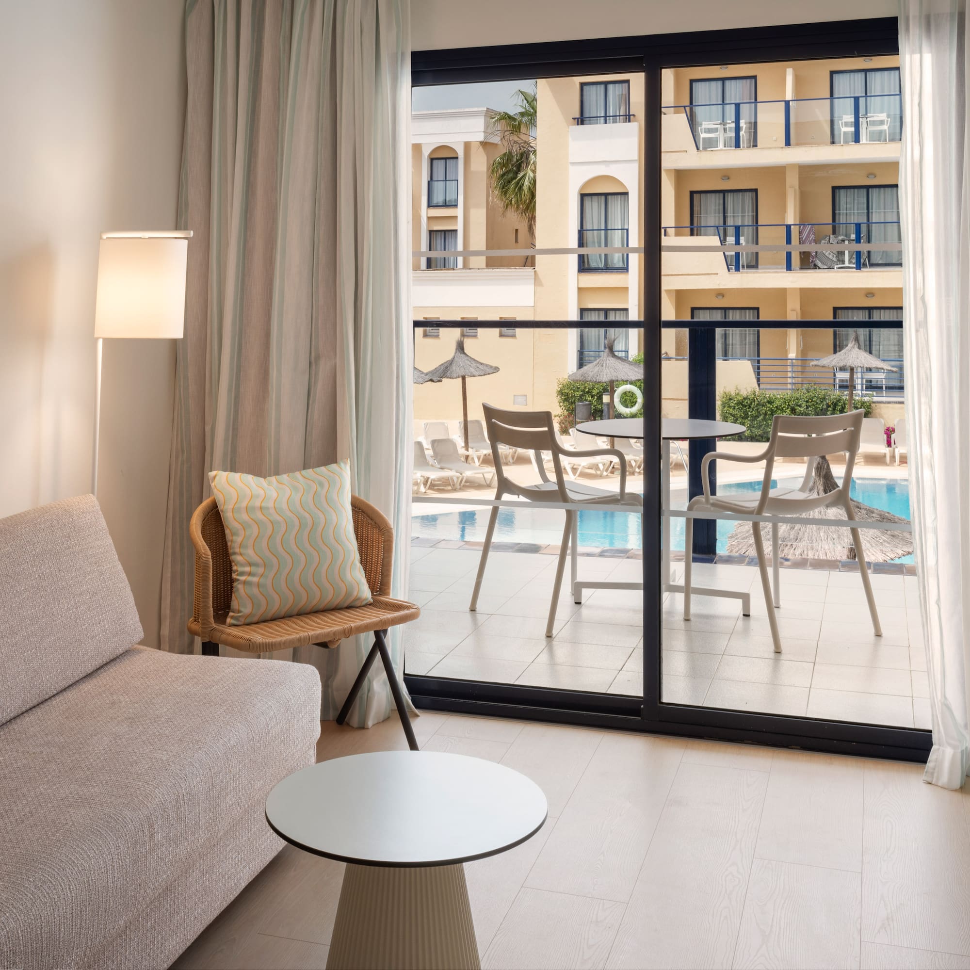 Modern hotel room with sofa, mirror, and balcony overlooking a resort pool.