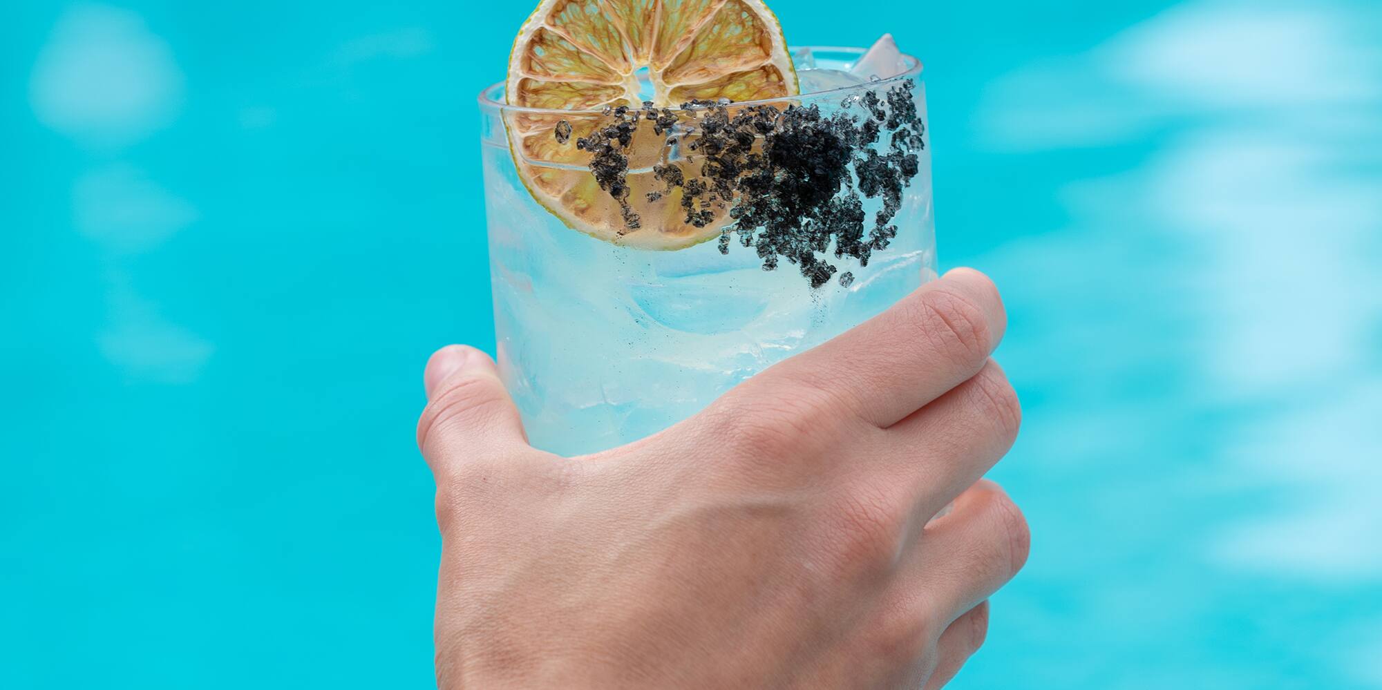a hand holding a glass of water with a slice of lemon and black poppy seeds