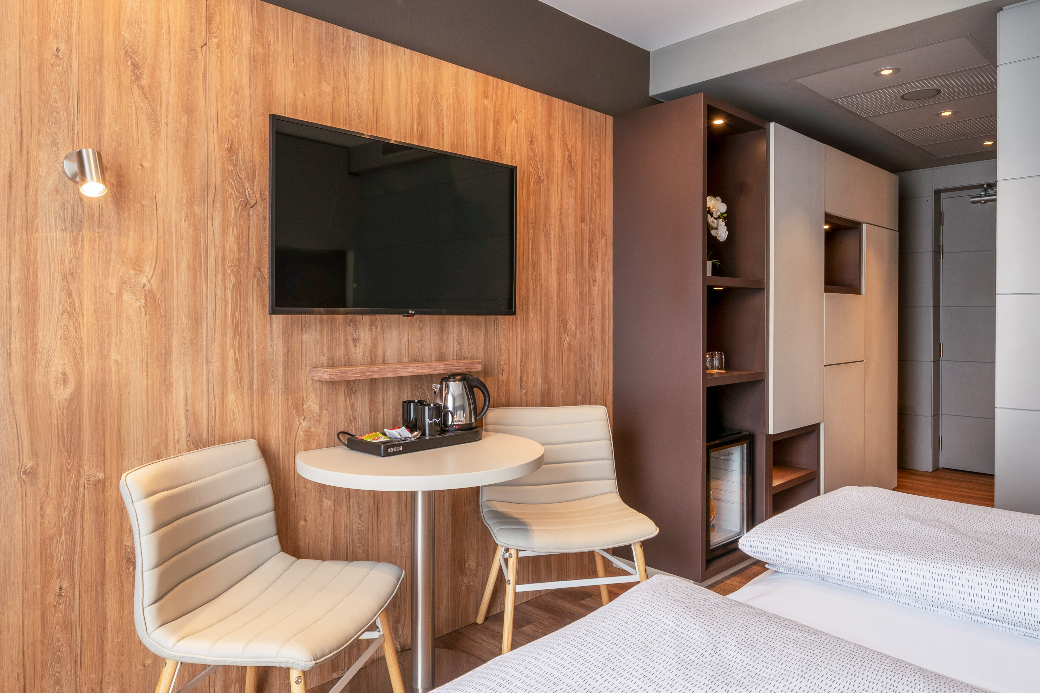 Modern hotel room featuring wood paneling, wall TV, coffee station, and seating.