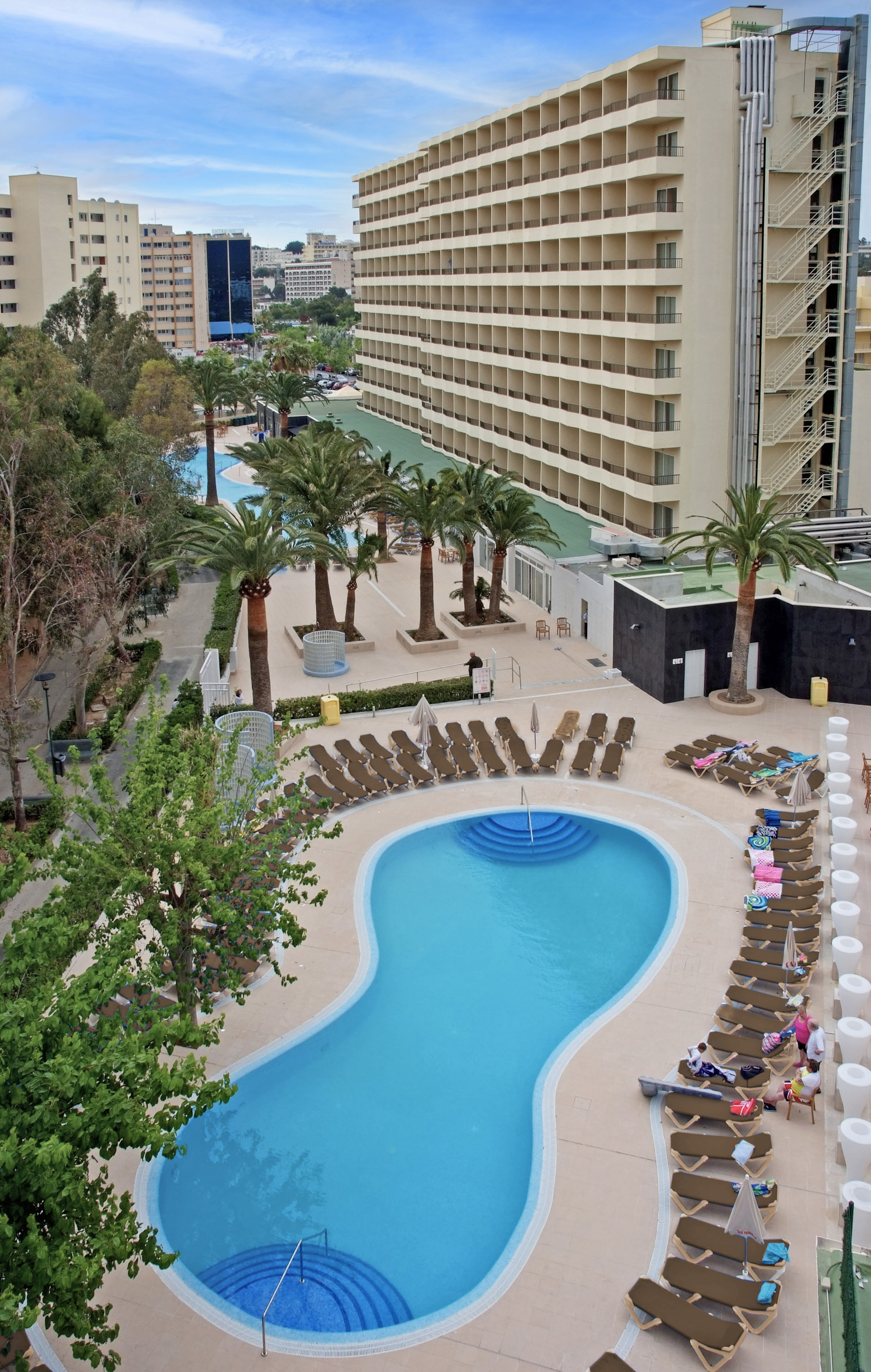 Resort swimming pool with sun loungers, palm trees, and hotel buildings under a cloudy sky.