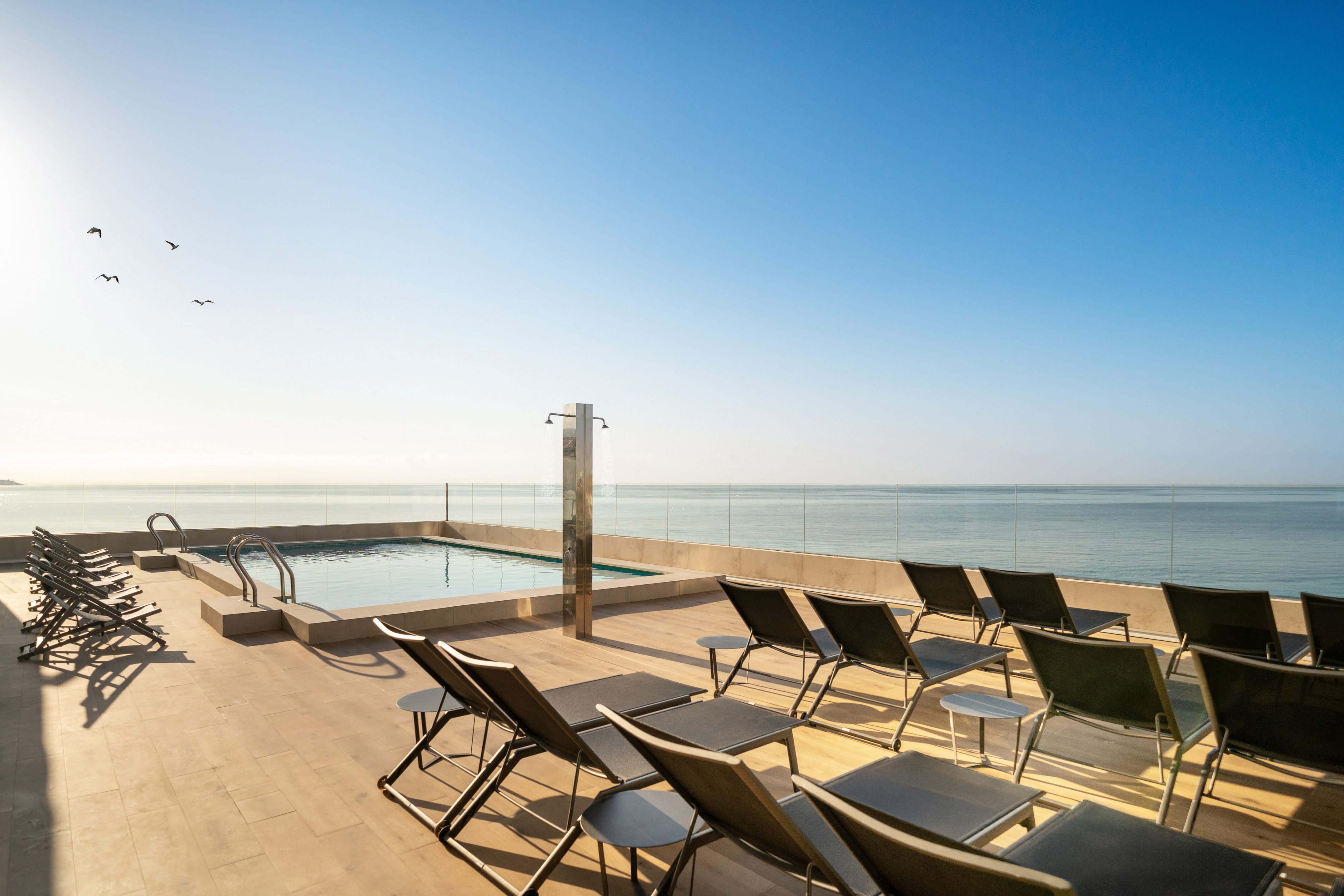 Luxury rooftop pool deck with ocean view, sun loungers, and clear sky.