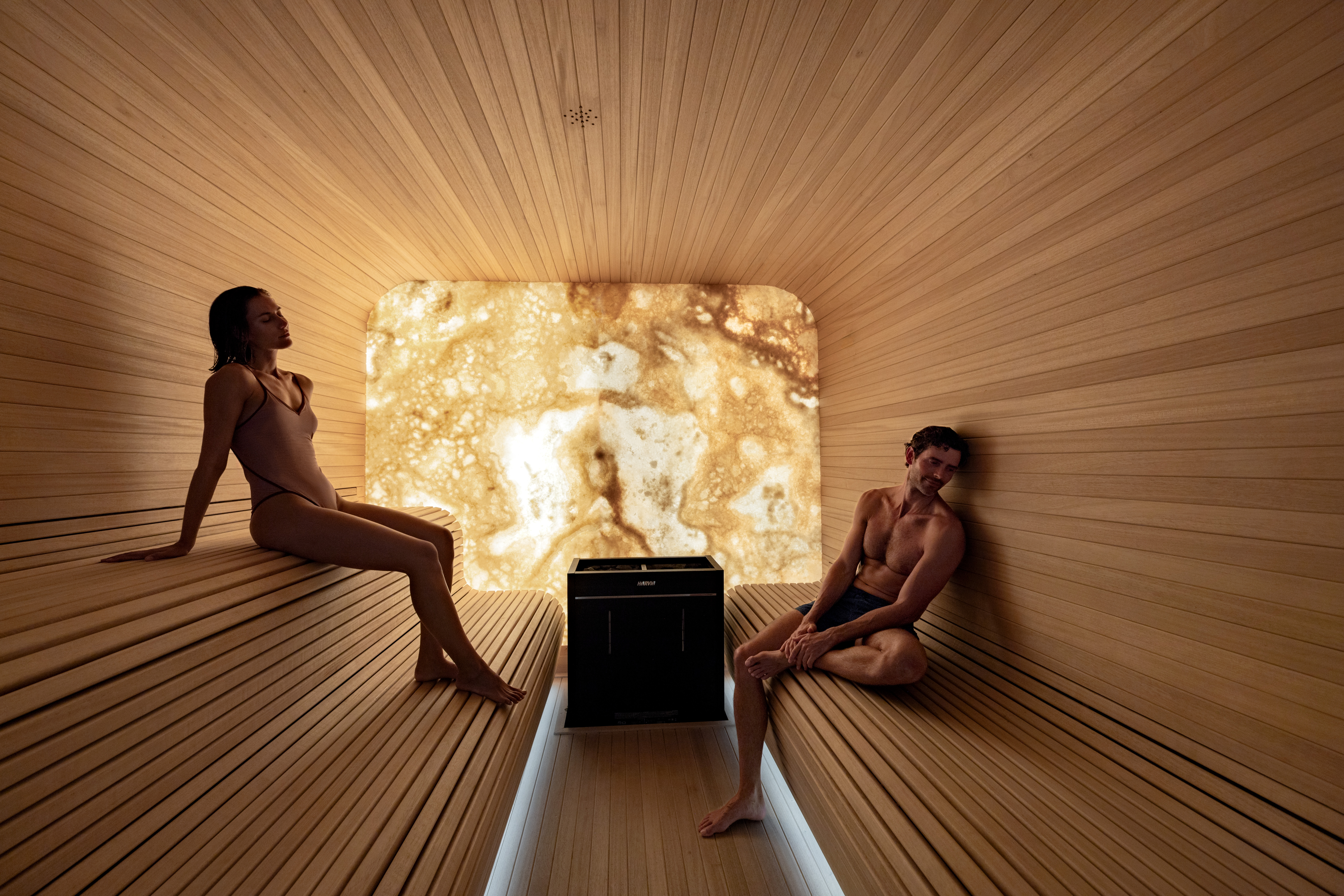 Man and woman relaxing in a sleek, modern wood sauna with illuminated back wall.