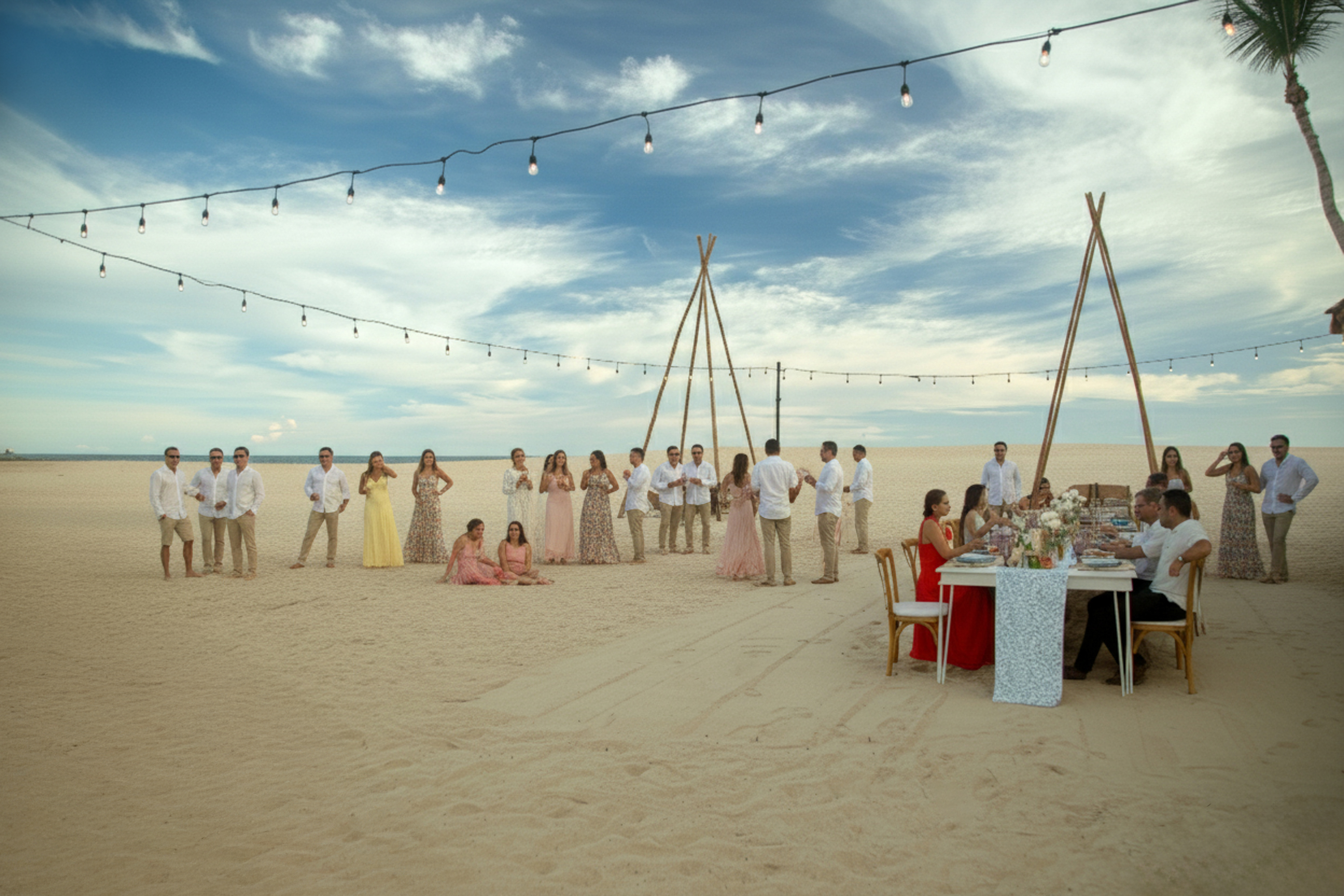 Beach wedding reception with guests, string lights, and dining table on sand.
