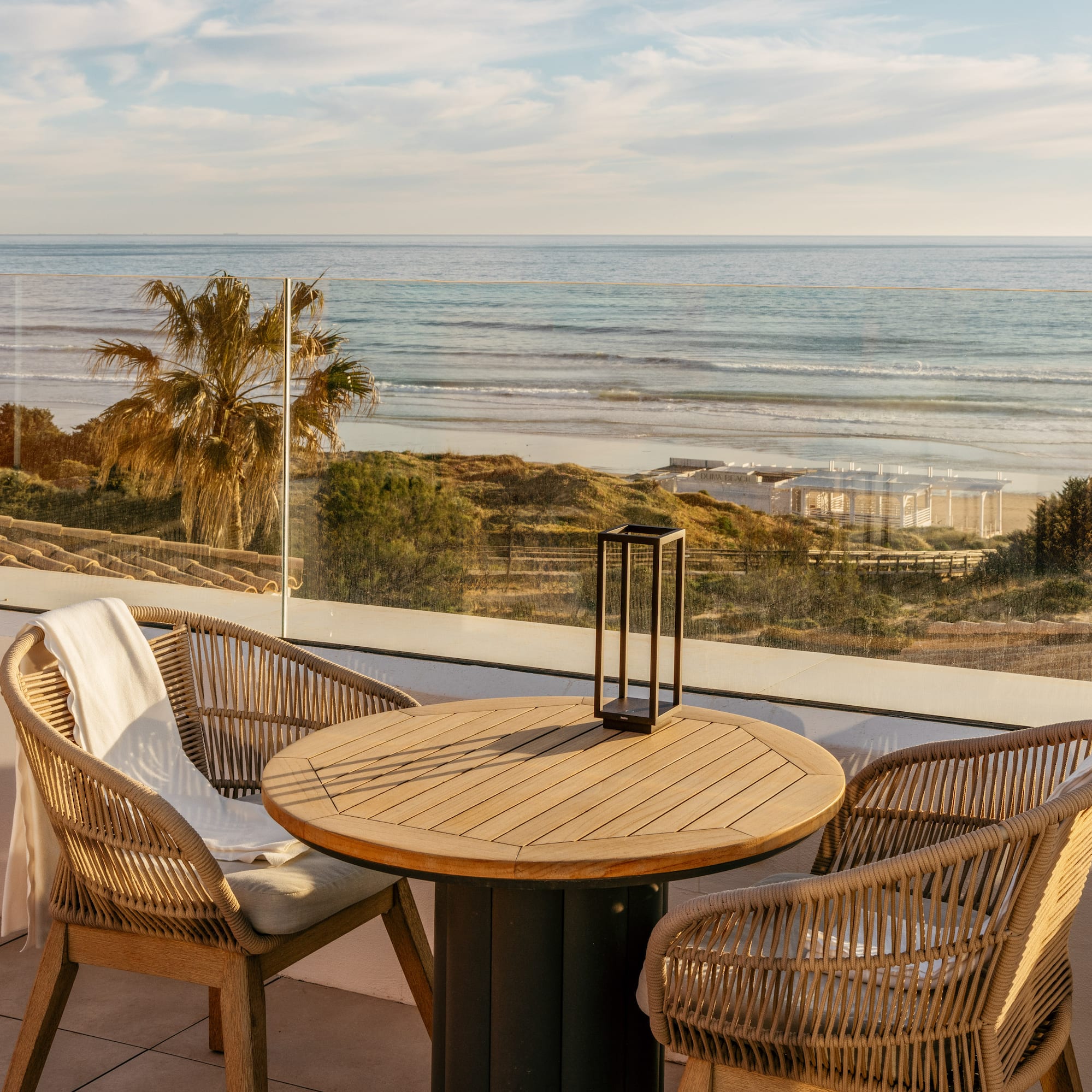 Sunny ocean view terrace with wicker chairs and wooden table by a sandy beach.