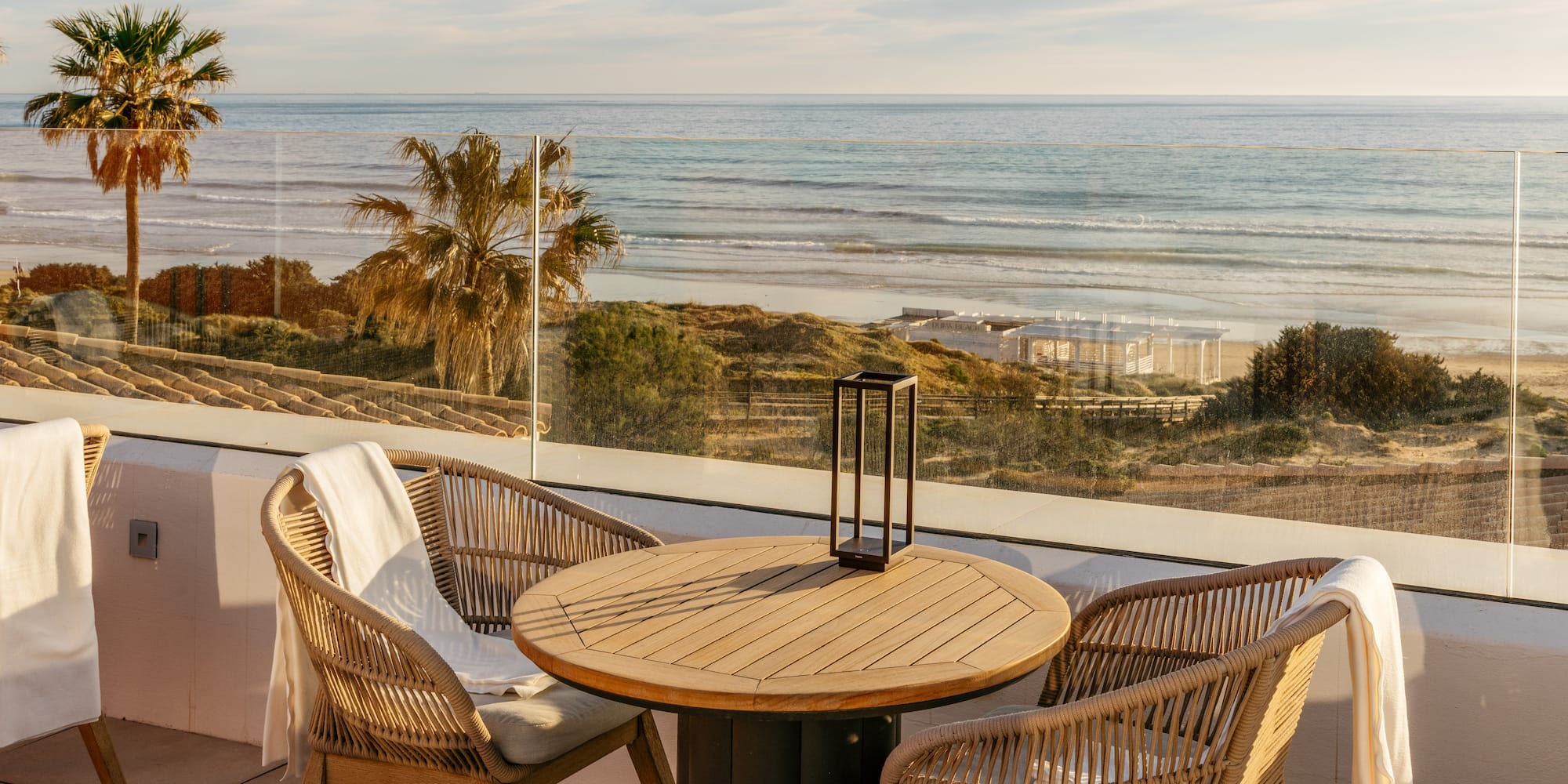 Coastal terrace with rattan chairs, wooden table, and ocean beach view, palm trees.
