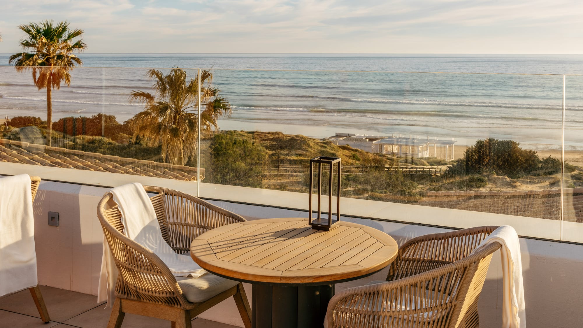 Coastal terrace with rattan chairs, wooden table, and ocean beach view, palm trees.