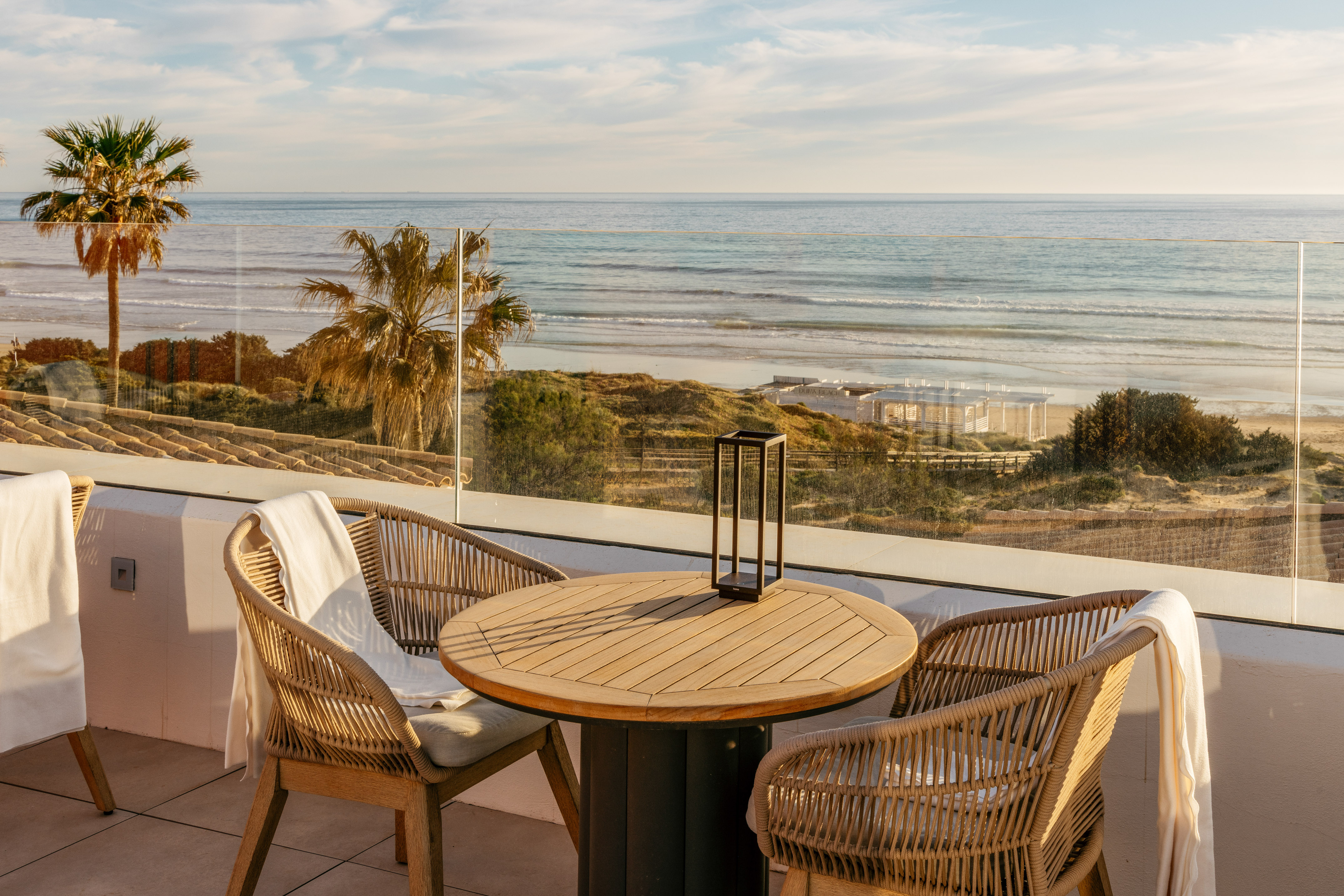 Coastal terrace with rattan chairs, wooden table, and ocean beach view, palm trees.