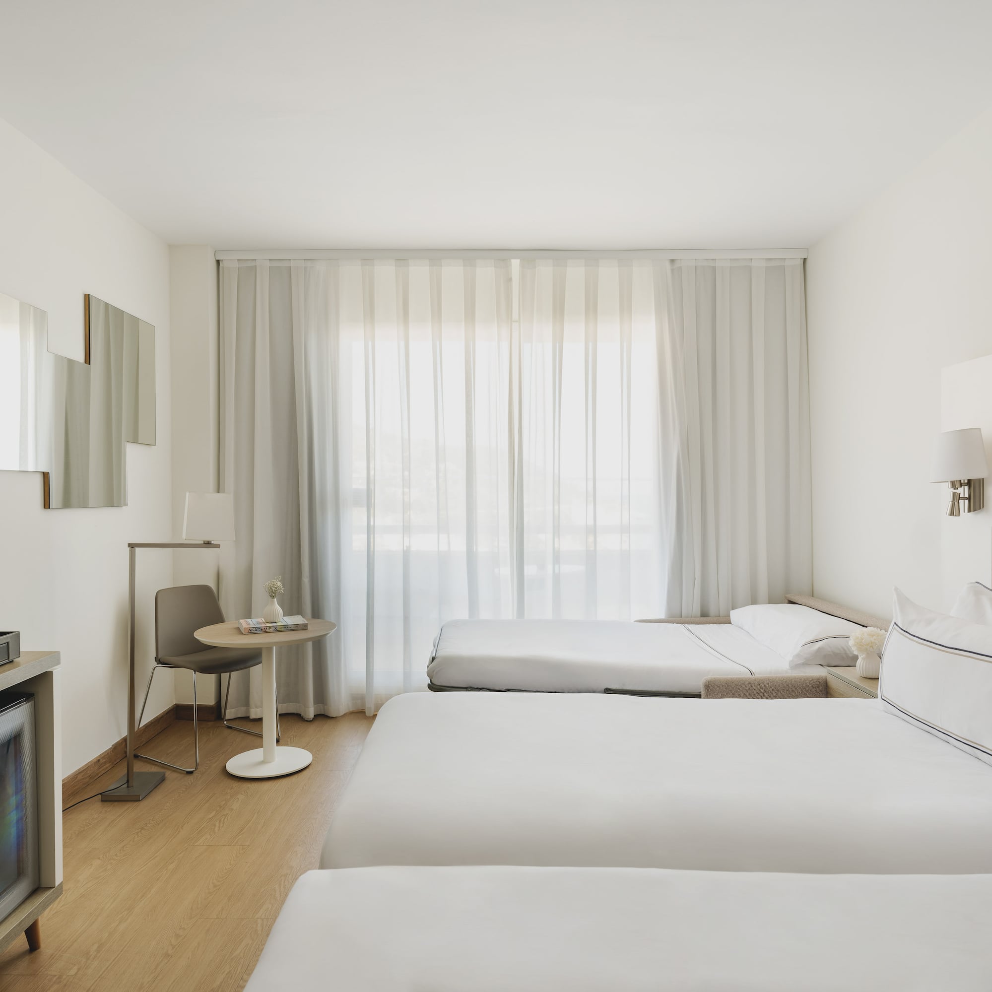 Habitación de hotel moderna y luminosa con tres camas, minibar y cortinas blancas.