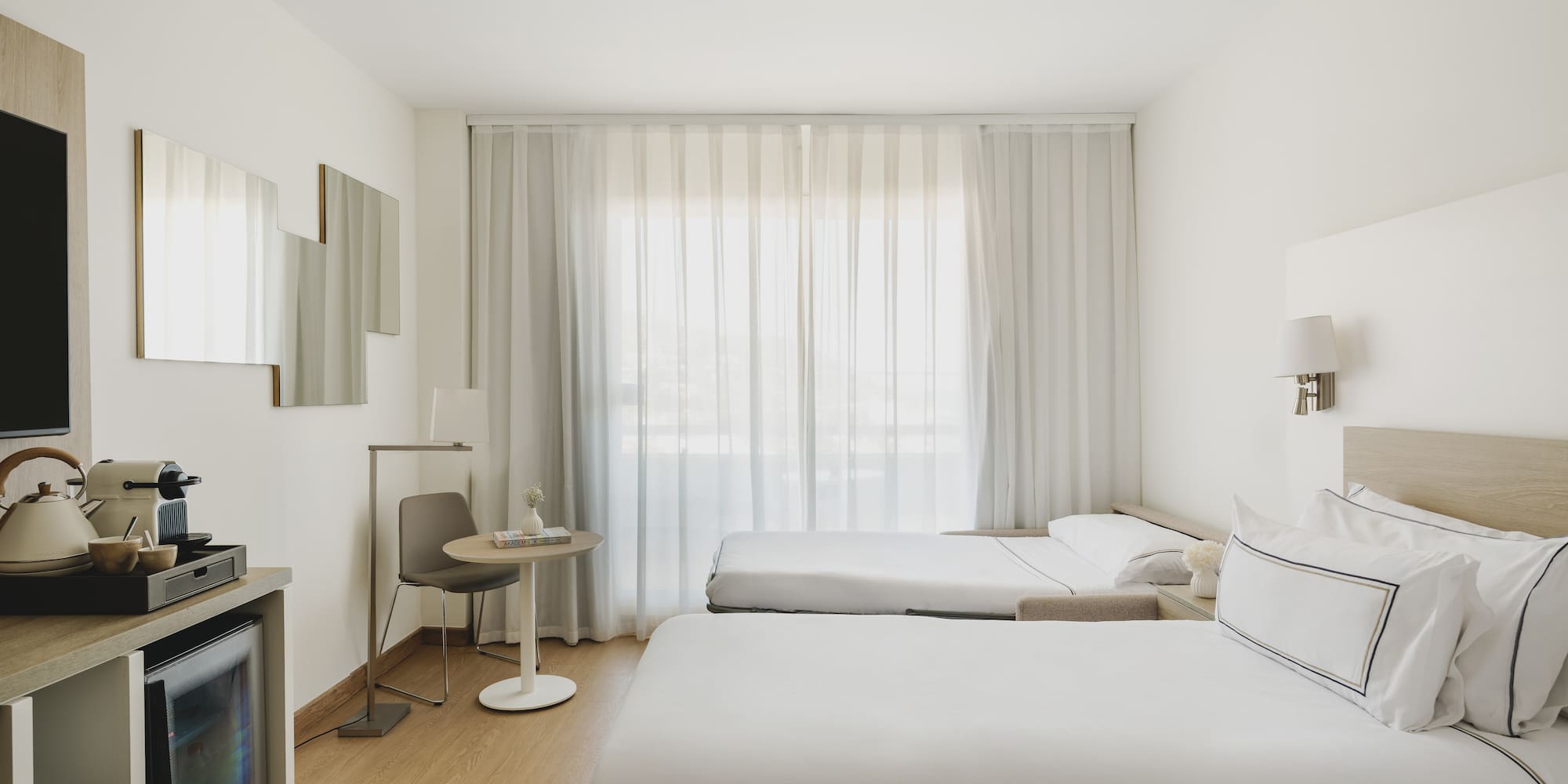 Habitación de hotel moderna y luminosa con tres camas, minibar y cortinas blancas.