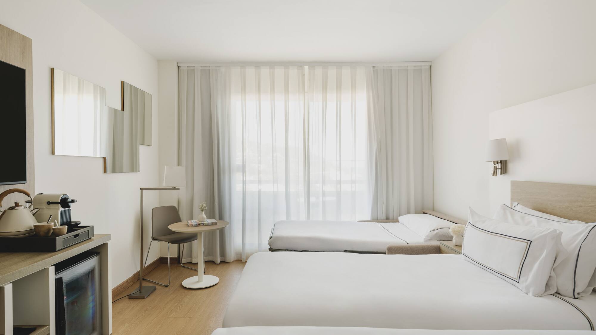 Habitación de hotel moderna y luminosa con tres camas, minibar y cortinas blancas.