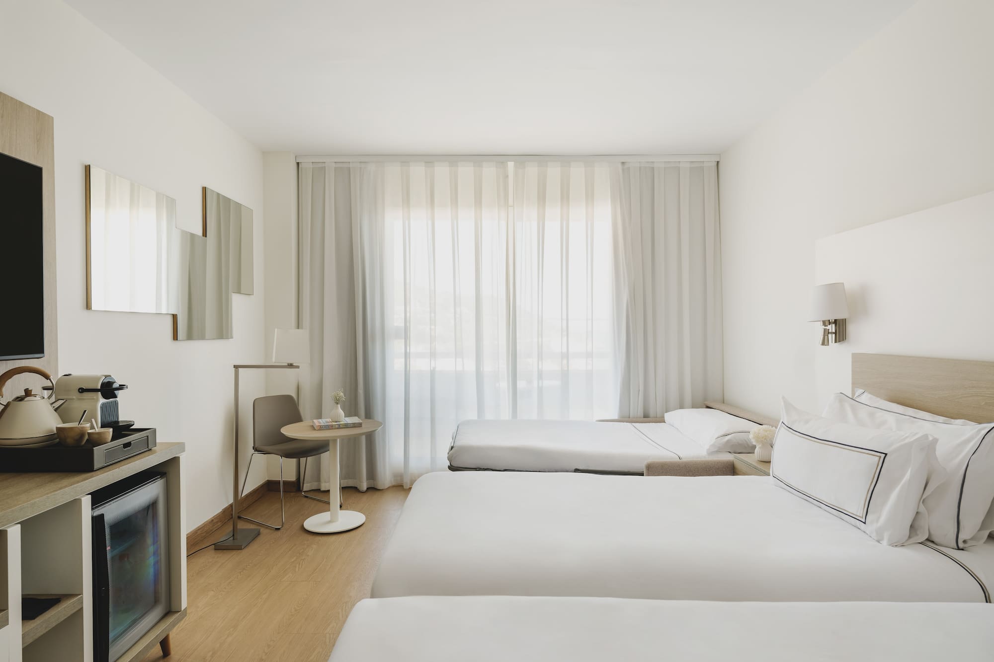 Habitación de hotel moderna y luminosa con tres camas, minibar y cortinas blancas.