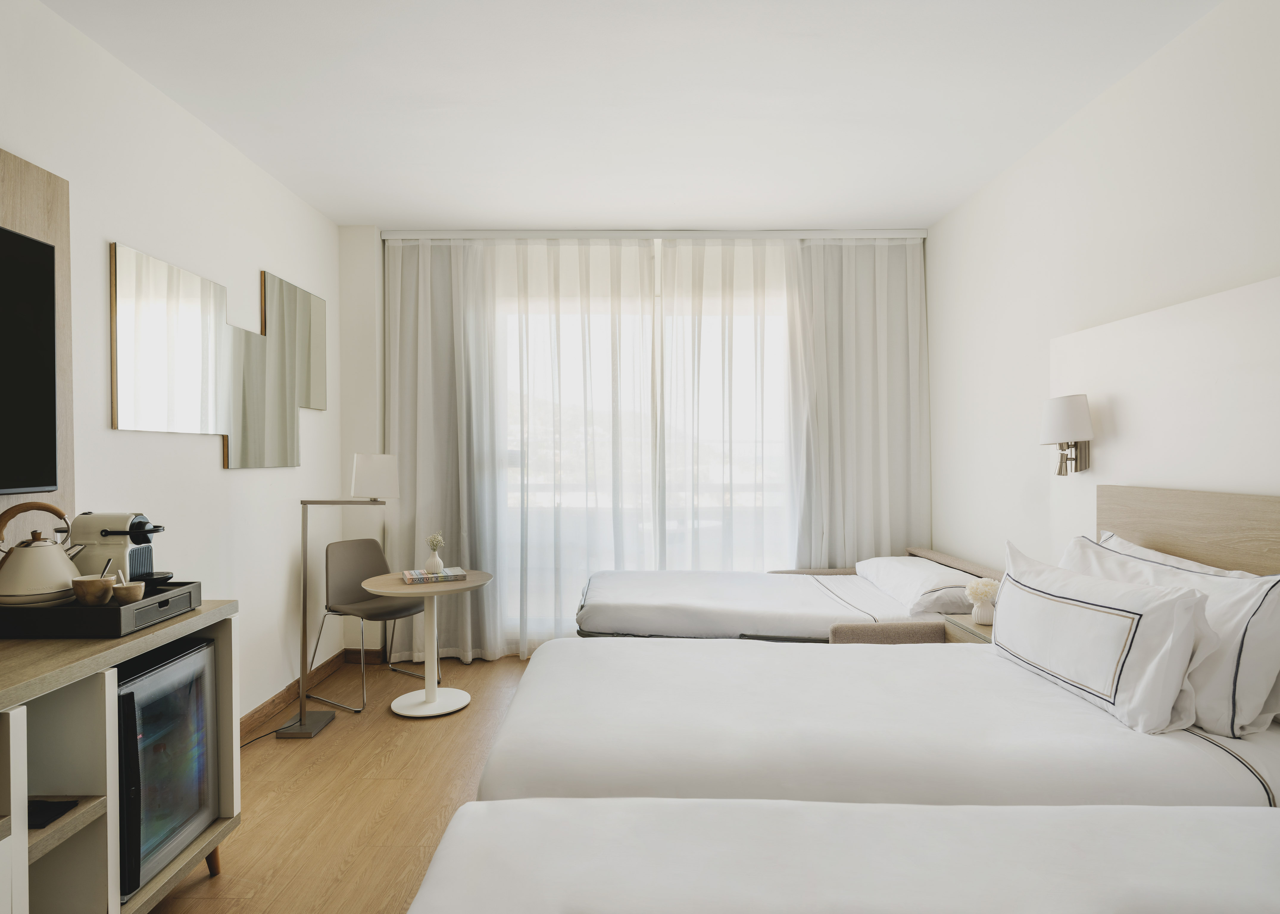 Habitación de hotel moderna y luminosa con tres camas, minibar y cortinas blancas.