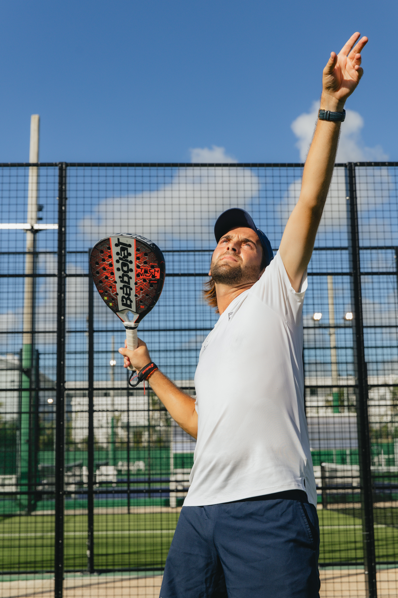 Male padel player holding Babolat racket on court, arm raised under blue sky.